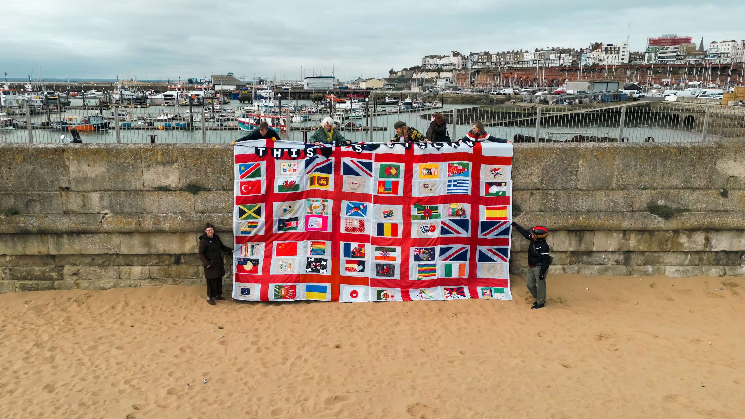 Seven people hold a large textile containing rows of decorated flag panels against a stone sea wall, with fishing boats and harbour buildings behind them.