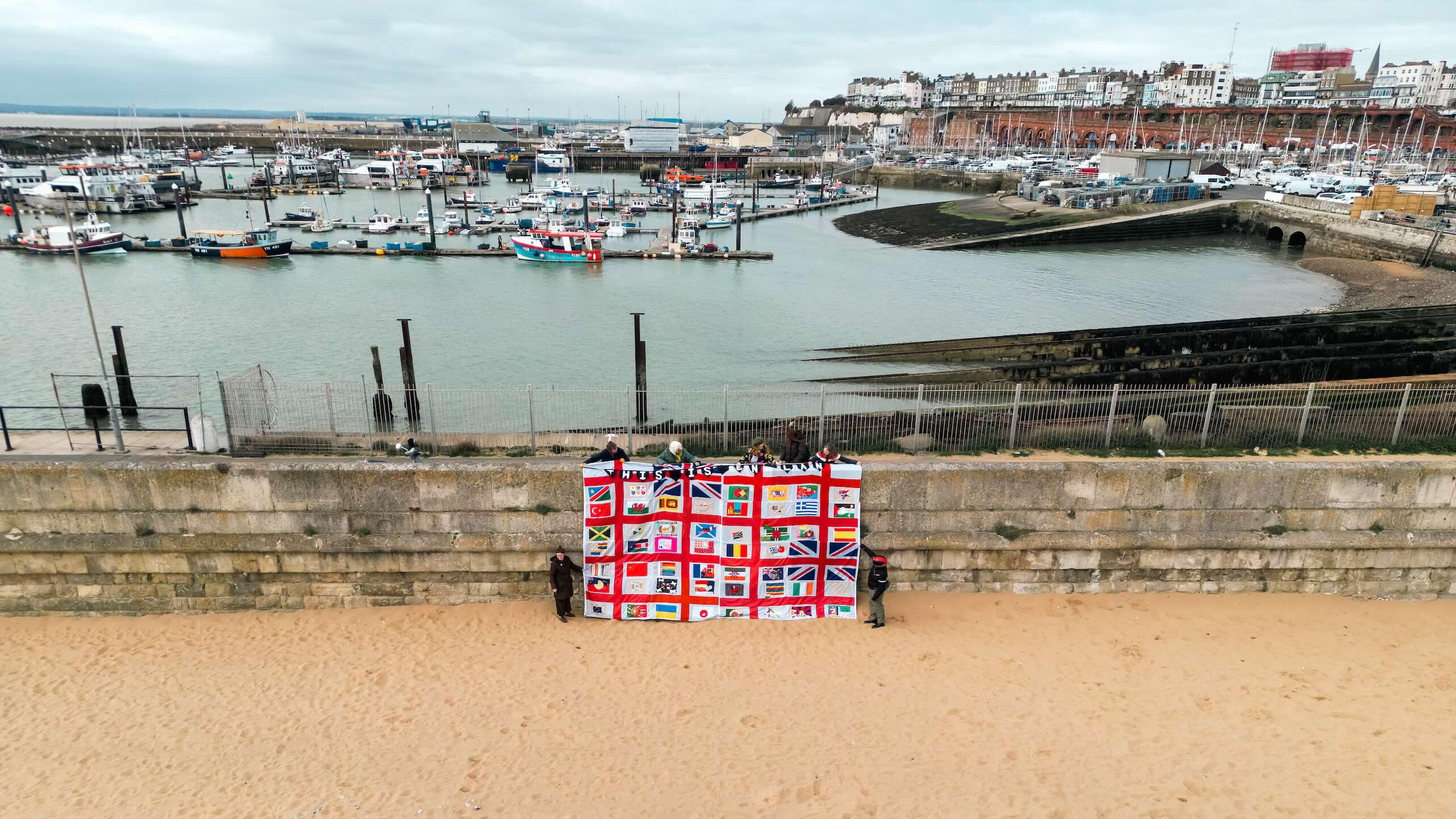 A high aerial view across Ramsgate Harbour, with the large patchwork textile and several people appearing small at the base of the sea wall.