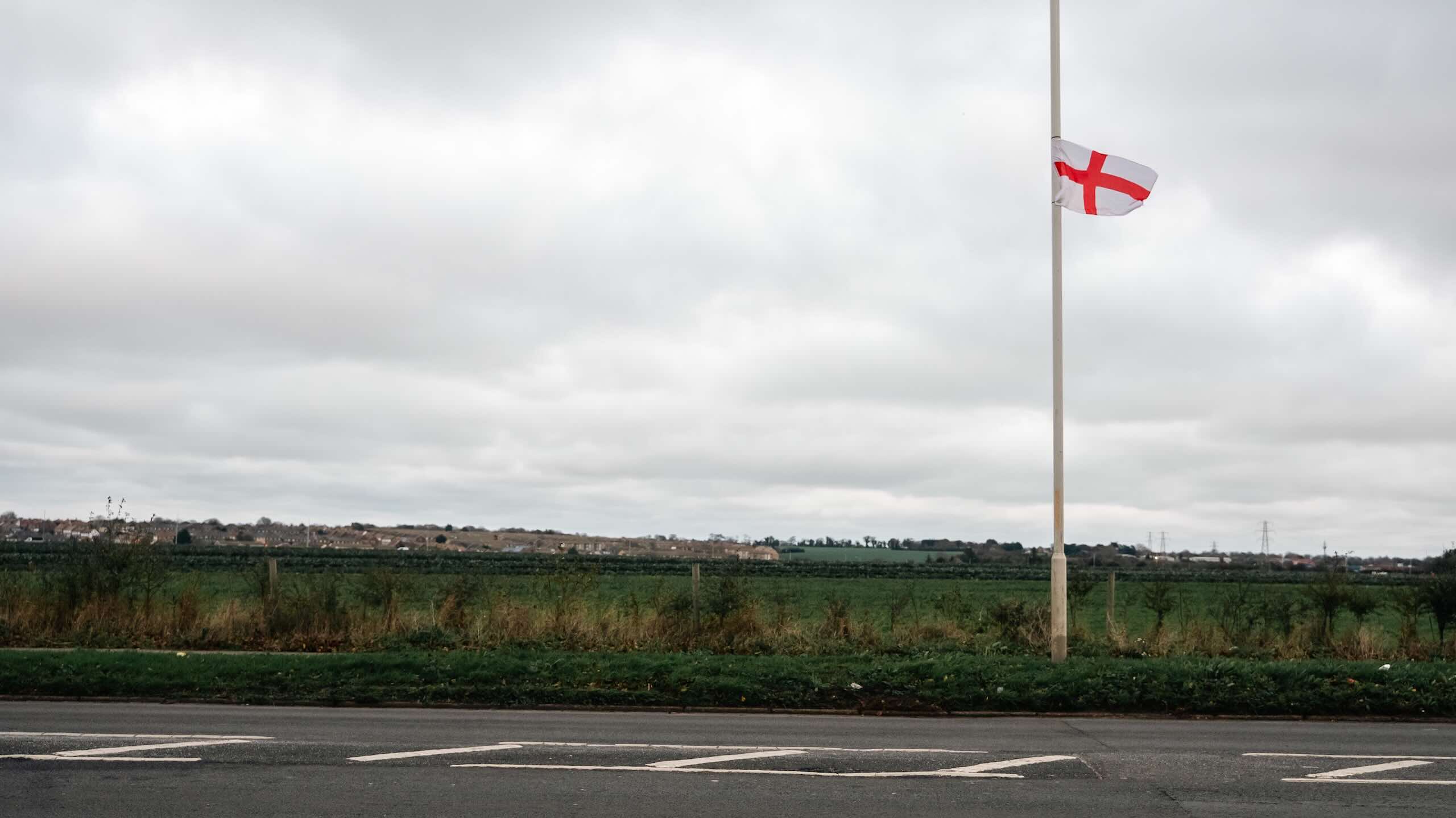 A small St George’s flag hangs partway down a street lamp beside a road, with green fields and an overcast sky behind it.