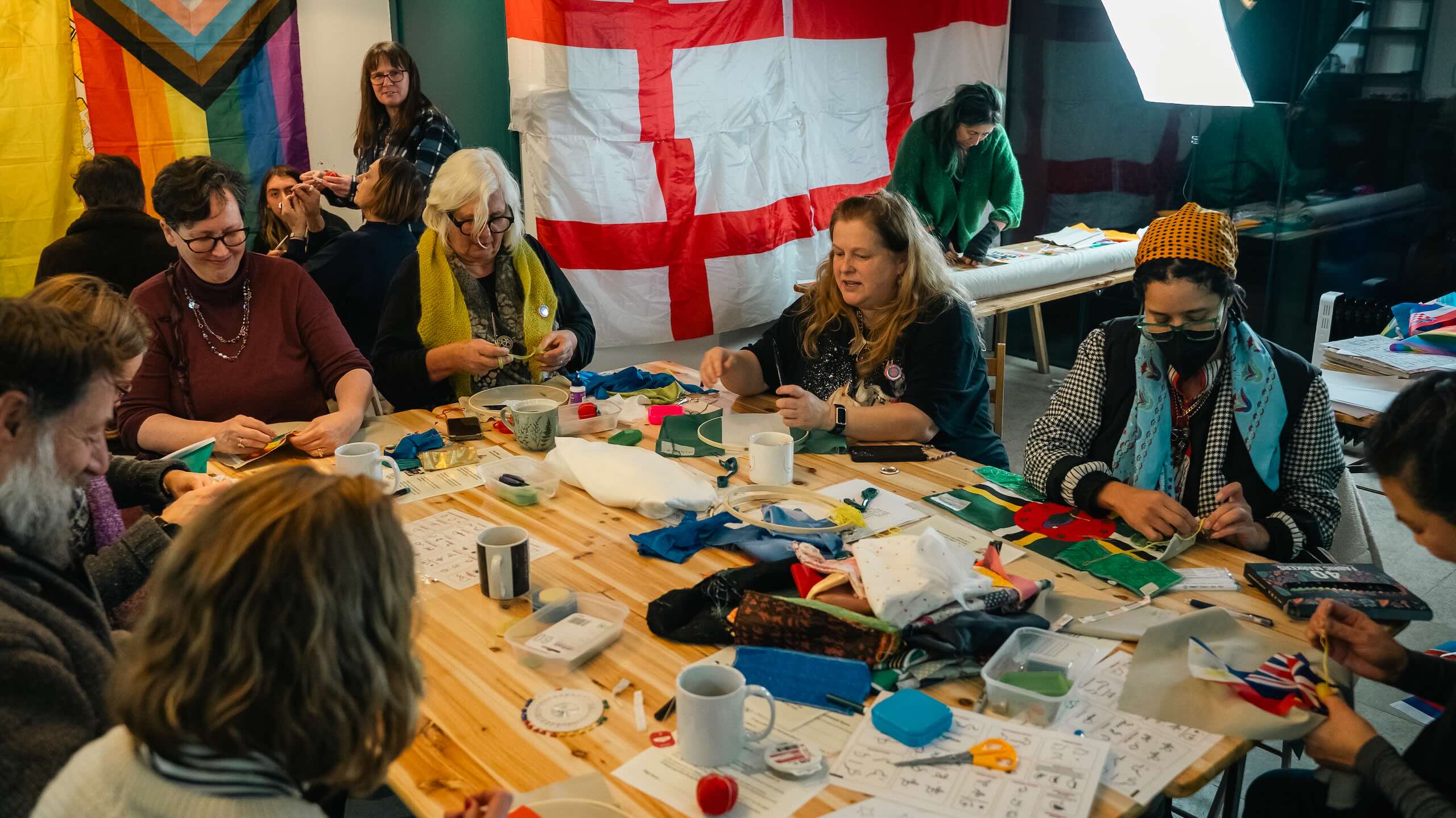 A large group sits around a wooden table covered with fabric, embroidery hoops, thread, scissors, mugs and sewing materials, with flags hanging behind them.