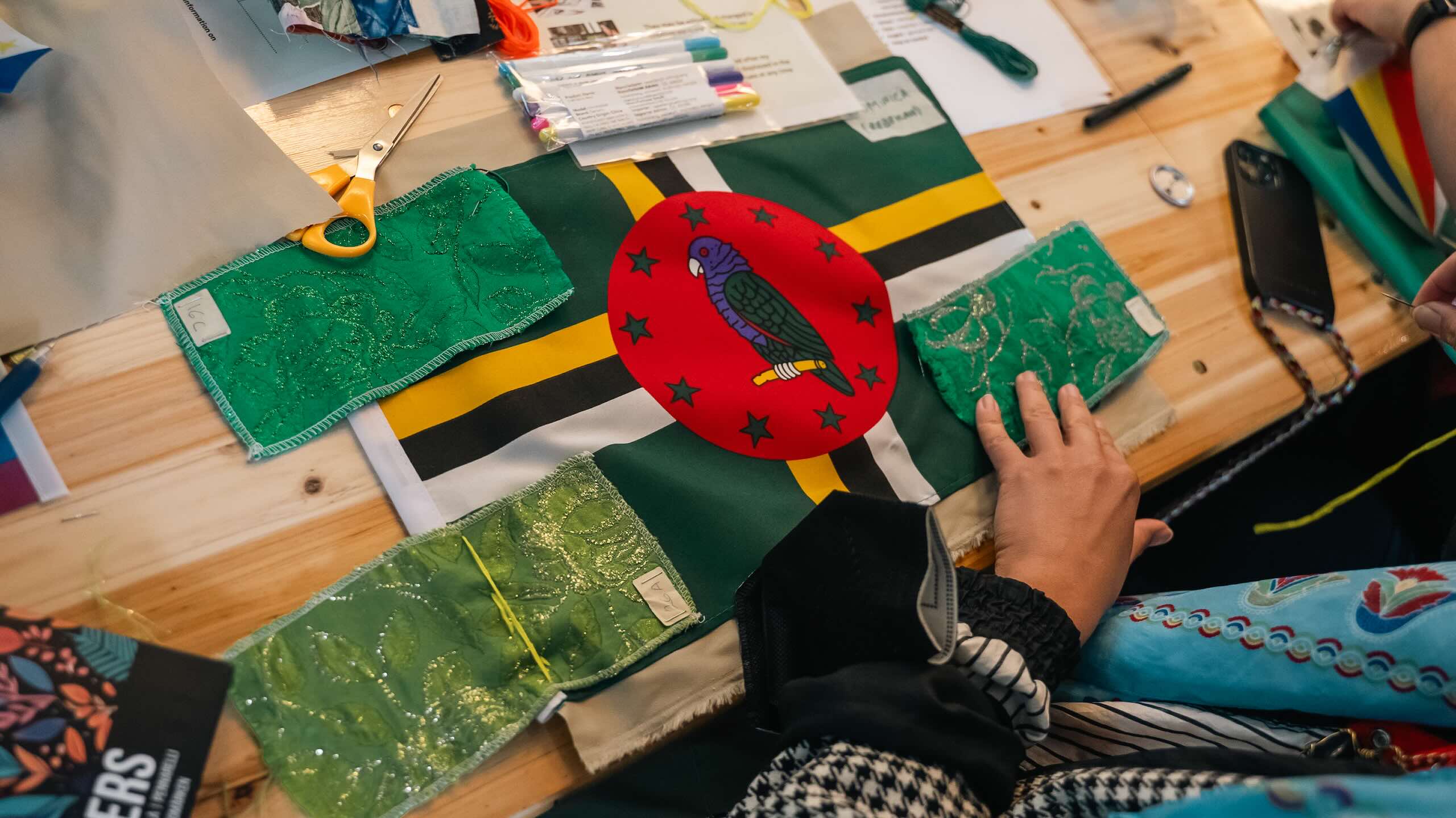 Hands arrange green patterned fabric pieces around a Dominica flag laid on a wooden table beside scissors, thread and pens.