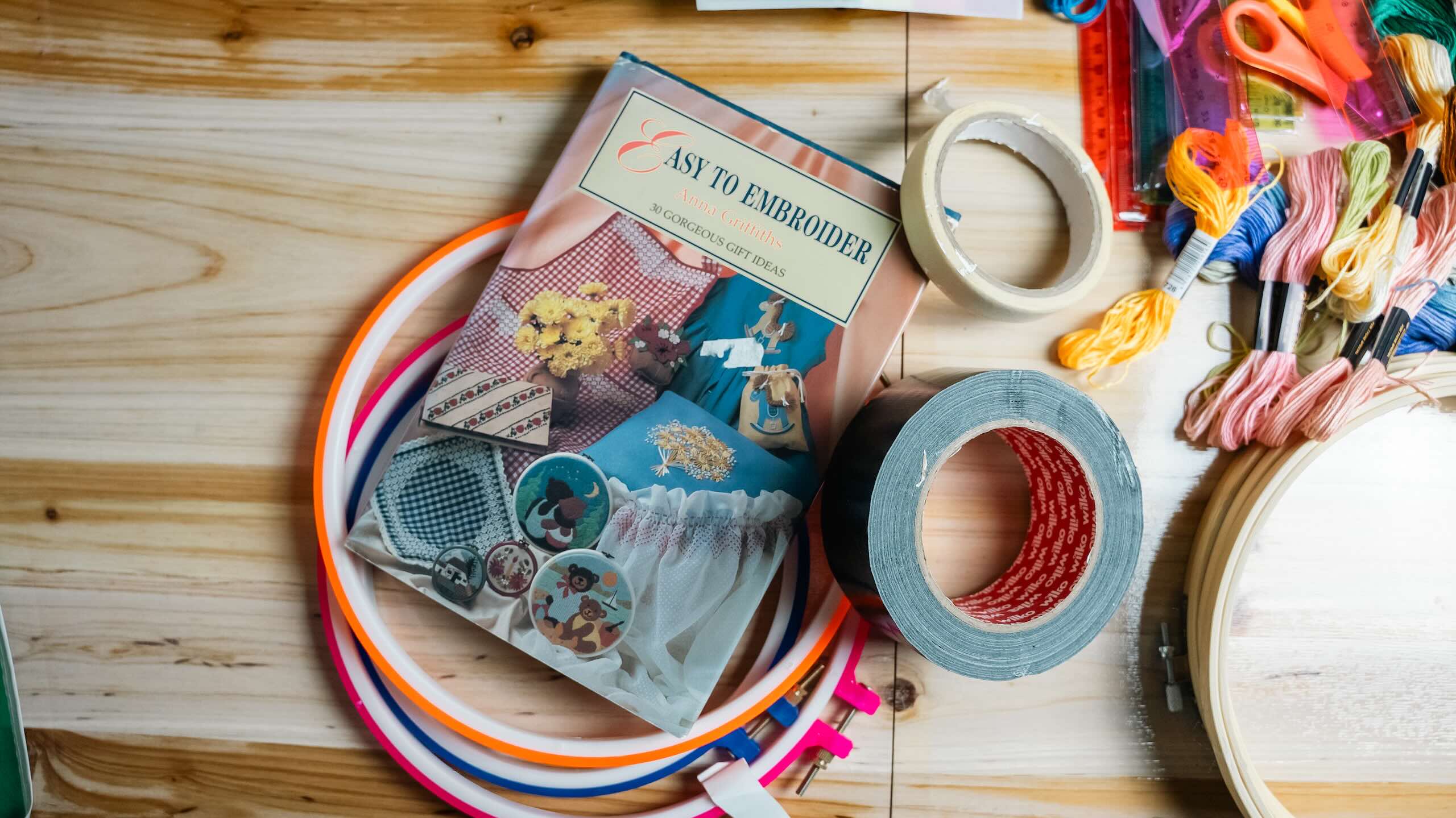 An overhead view of colourful embroidery hoops, embroidery thread, scissors, tape and an embroidery book arranged on a wooden table.