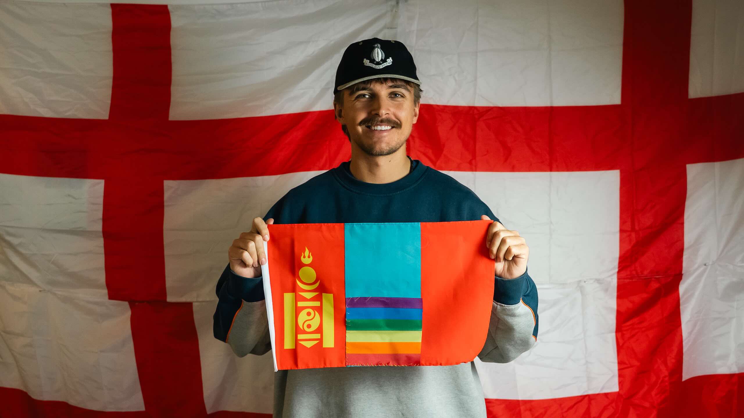 A smiling person wearing a dark cap holds a red and blue textile panel featuring the Mongolian emblem and horizontal rainbow-coloured strips.