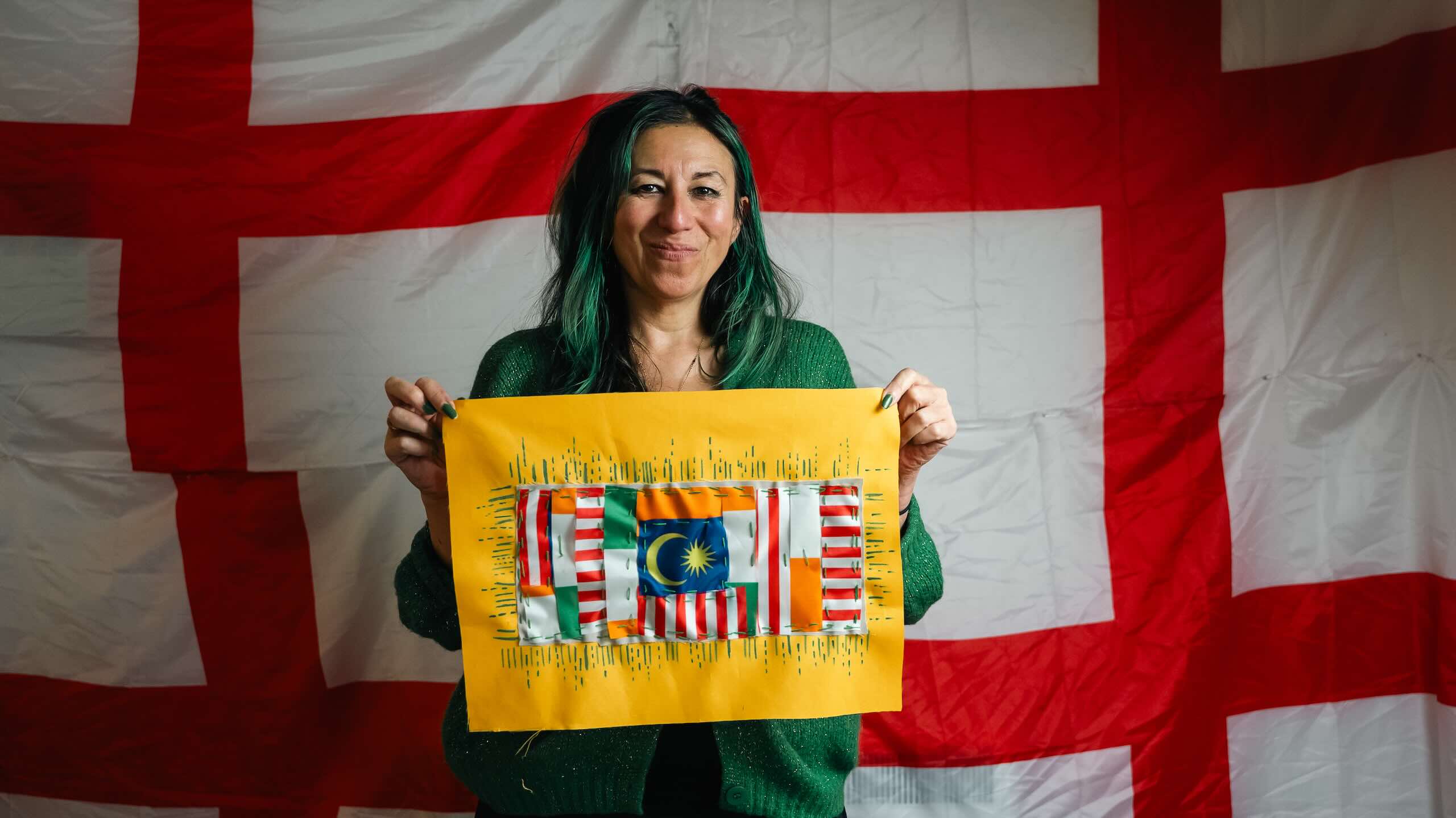 A person with green-streaked dark hair holds a yellow-backed textile artwork incorporating red, white, green and blue flag fabrics.