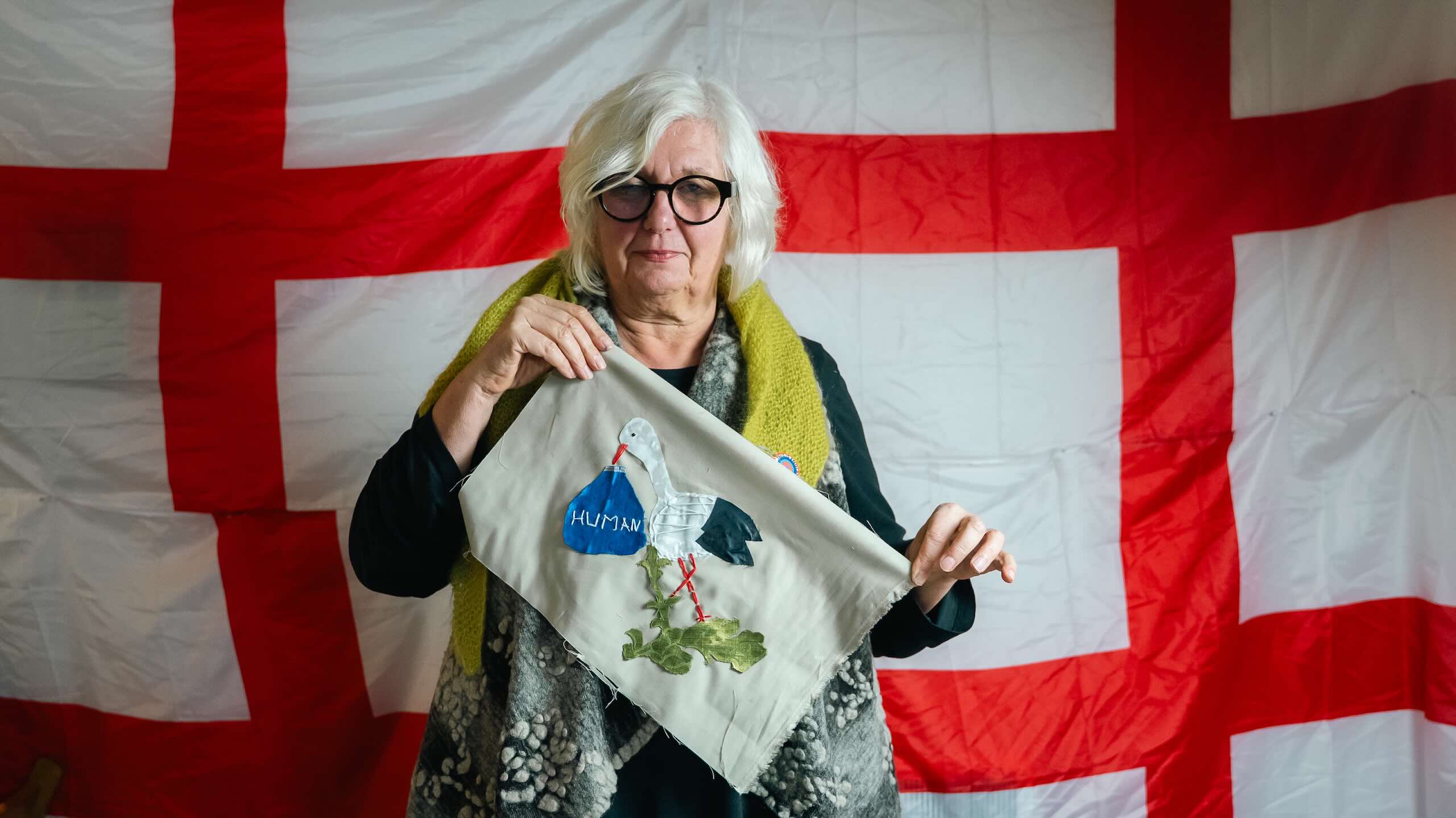 An older person with white hair and black glasses holds a beige fabric panel featuring a white bird, green land shapes and the word “HUMAN.”