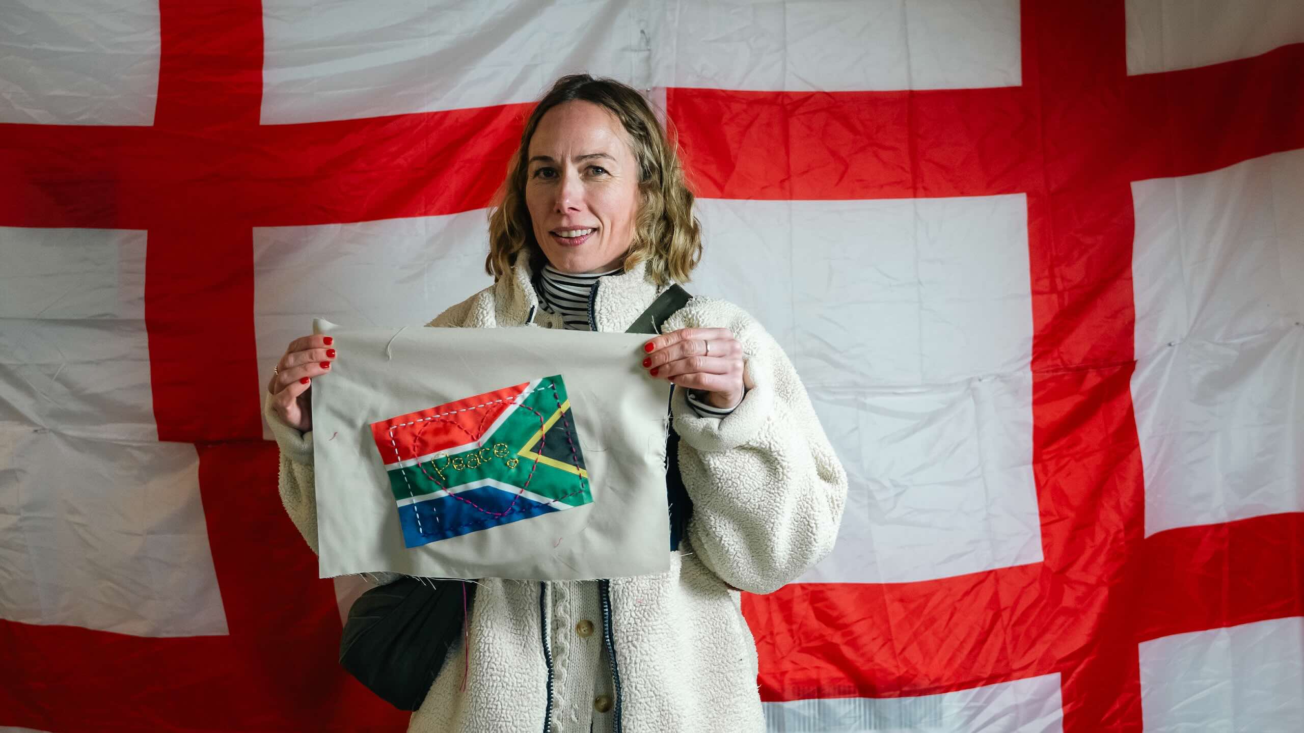 A smiling person in a cream fleece holds a beige fabric panel with a small South African flag crossed by colourful lines of hand stitching.