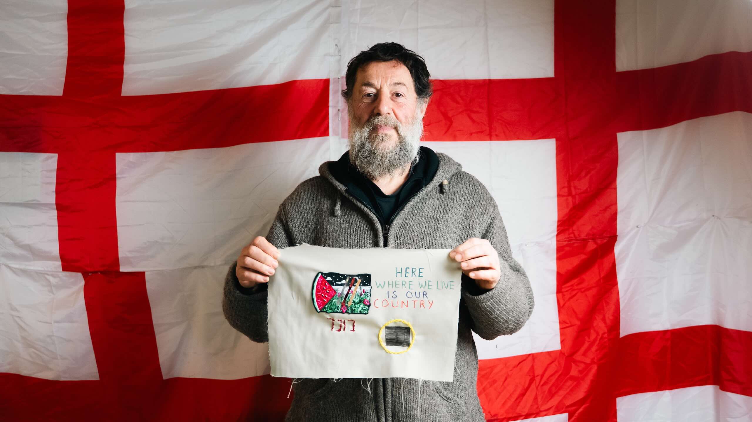 A bearded person holds a beige textile panel showing an embroidered landscape and watermelon with the words “HERE WHERE WE LIVE IS OUR COUNTRY.”