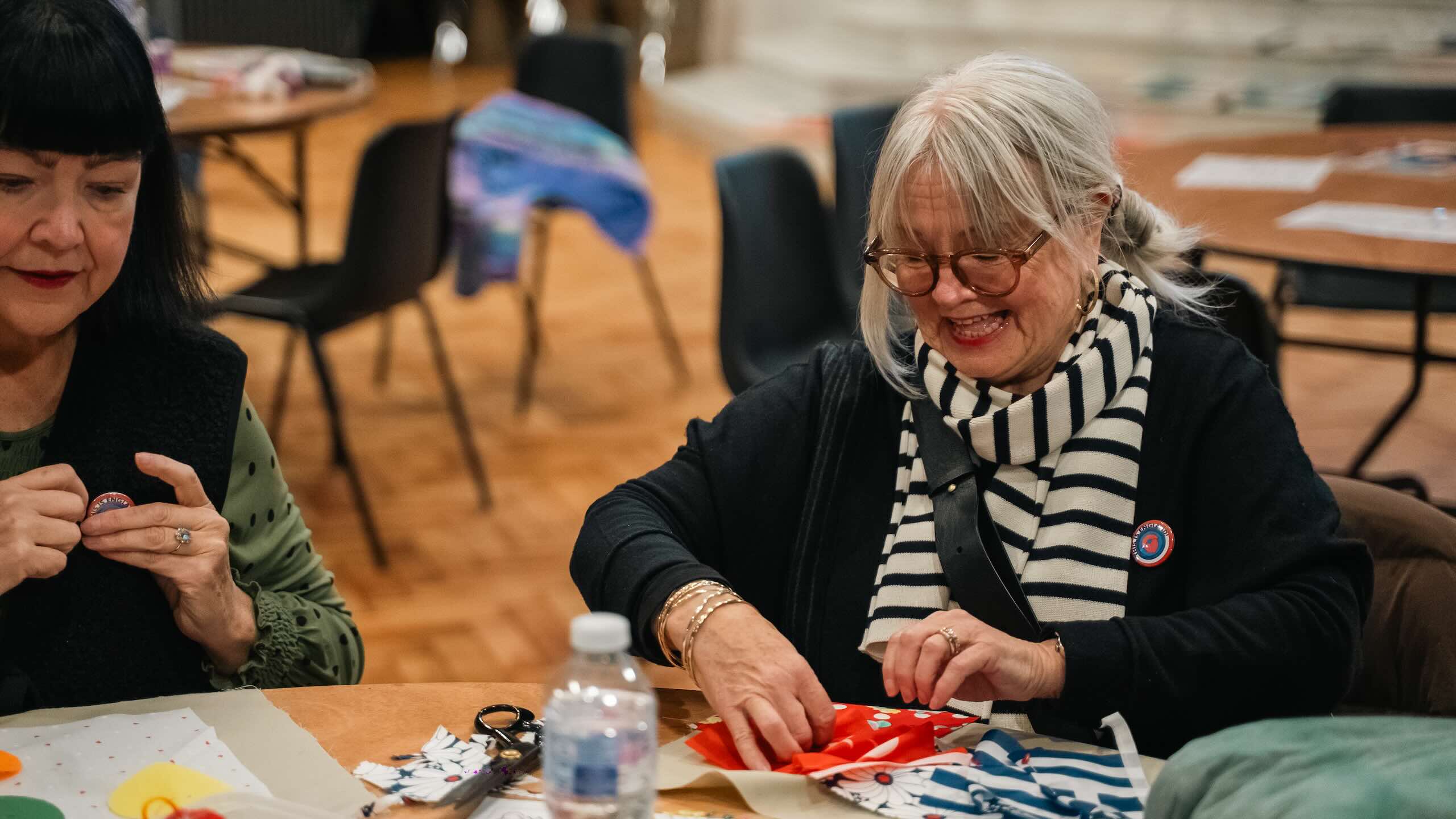 Two older people sit at a table working with red, blue, white and patterned fabric pieces, scissors and sewing materials.