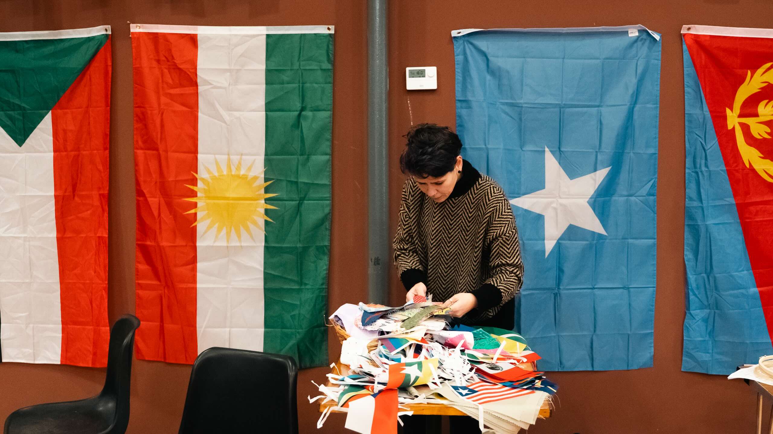 A person stands behind a table piled with cut flags and fabric scraps, with large Kurdistan, Somalia and Eritrea flags hanging on the wall.