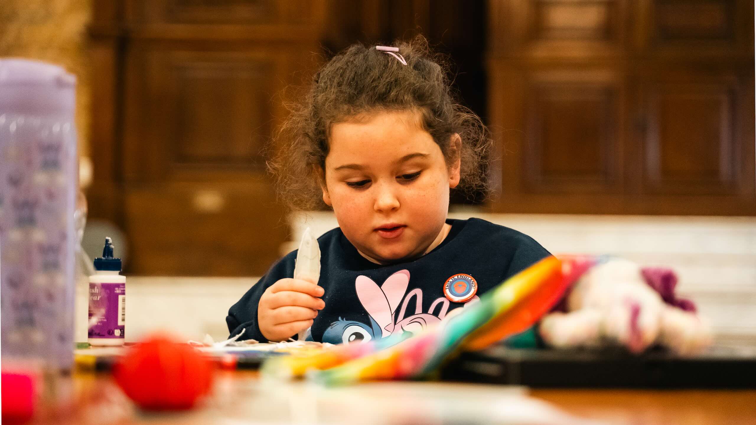 A child sits at a table concentrating on a craft activity, holding a pale feather or fabric shape among glue, colourful materials and sewing supplies.