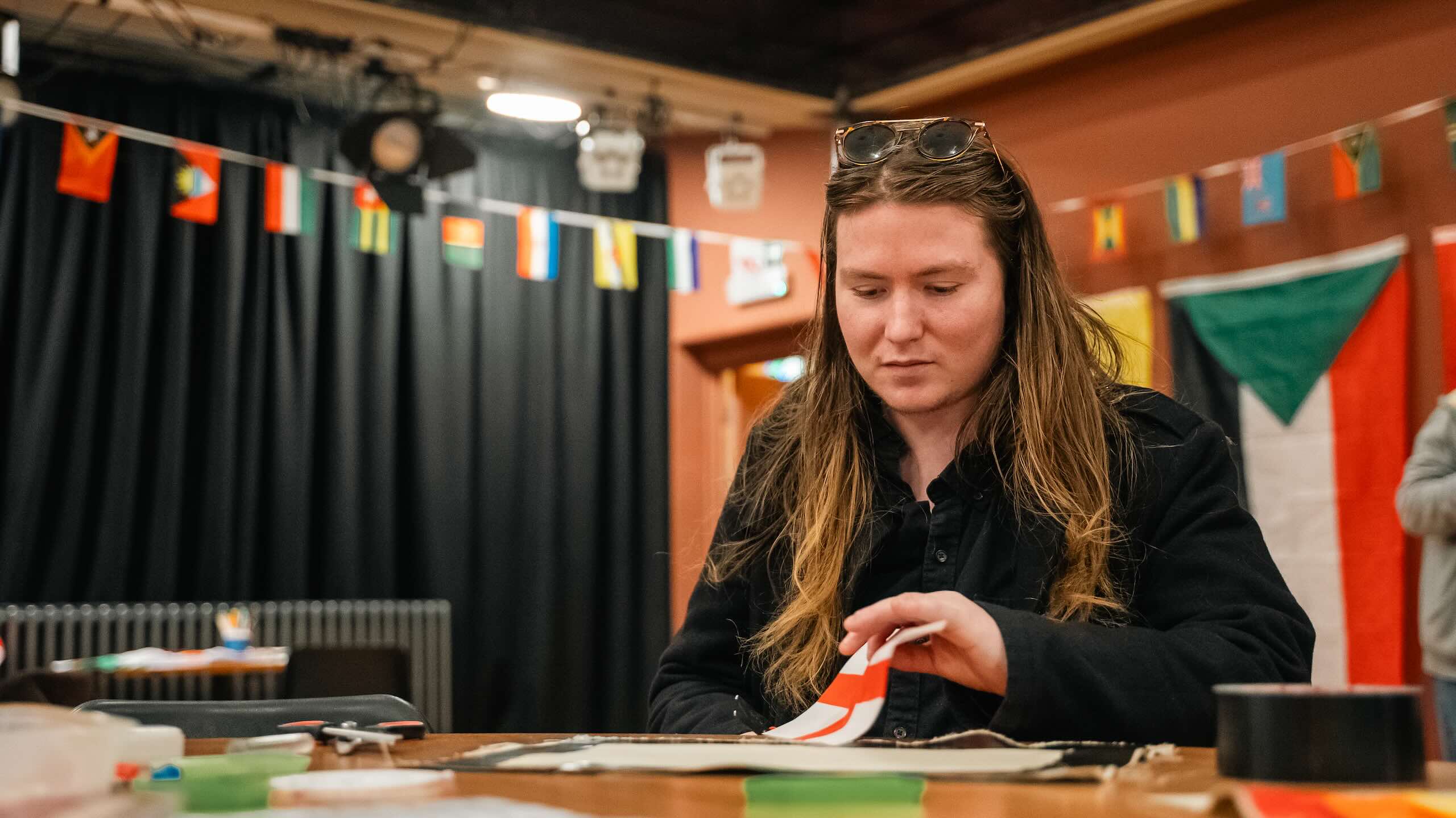 A person with long hair works at a table with fabric and scissors, with a line of small international flags and larger flags hanging behind them.