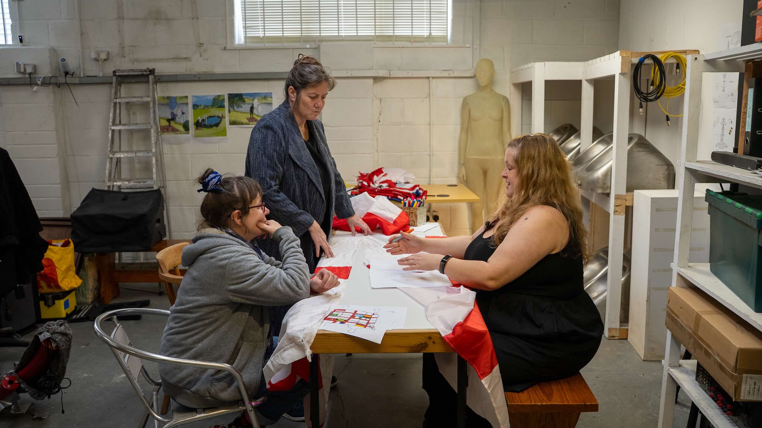 Three people gather around a worktable covered with red-and-white flag fabric, paper plans and pens in a studio space.