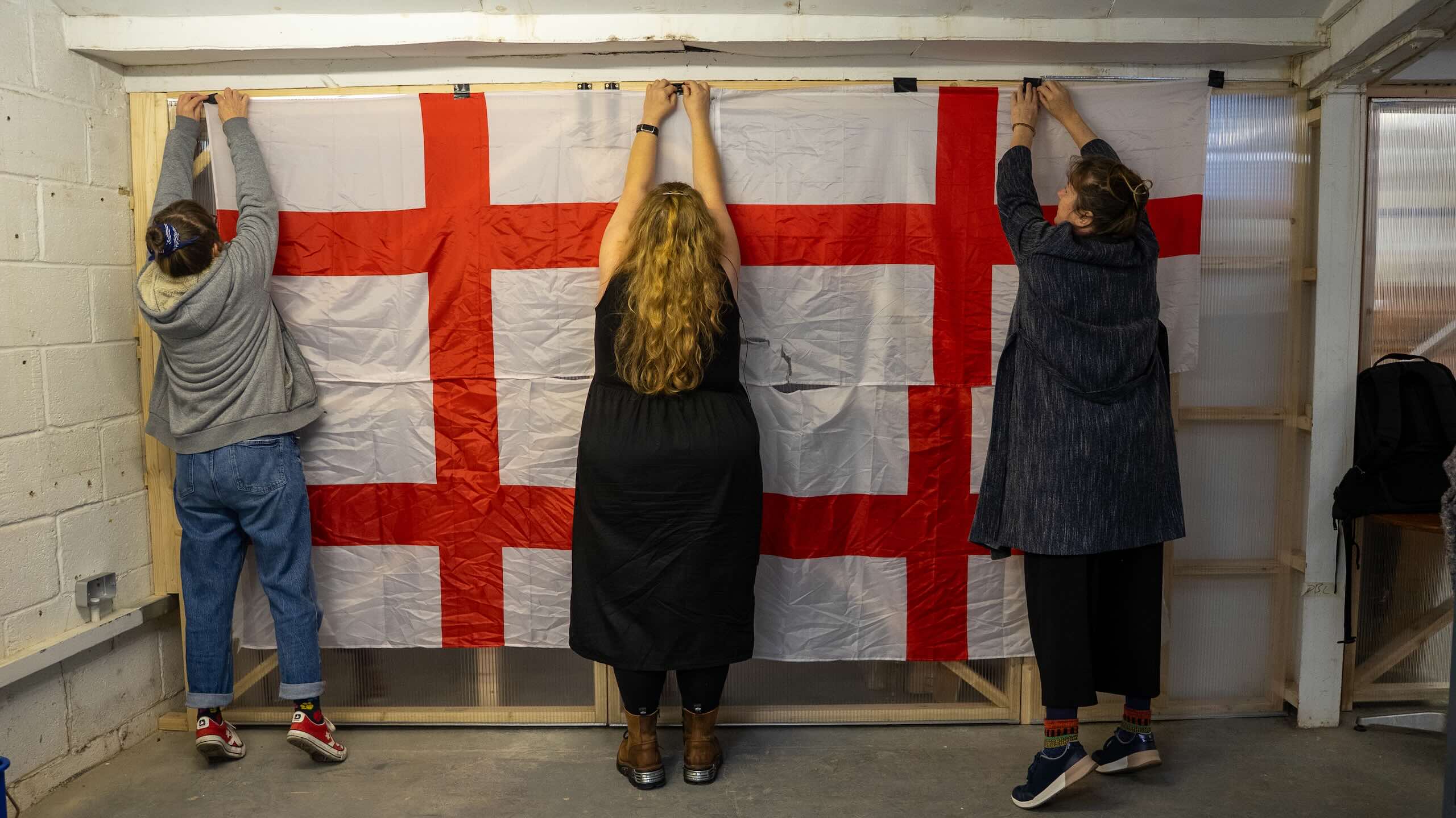 Three people stand with their backs to the camera, reaching upwards to fasten a large red-and-white cross-patterned textile to a wooden frame.
