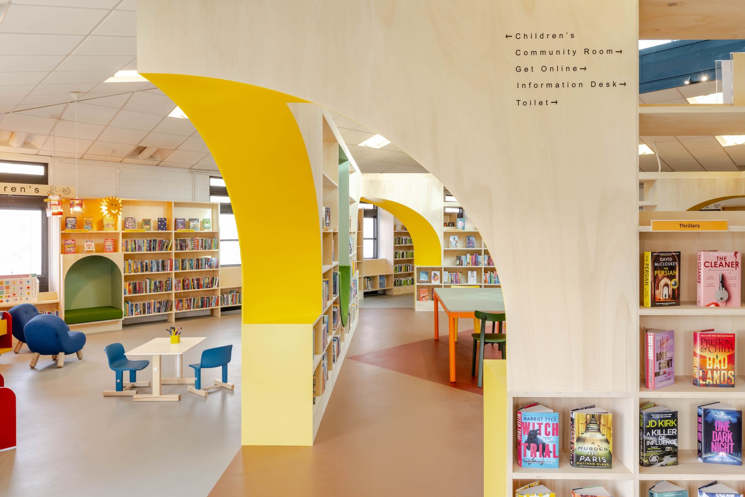 Interior view showing yellow archways, directional signage, bookshelves and children's seating area within Haydock Library.