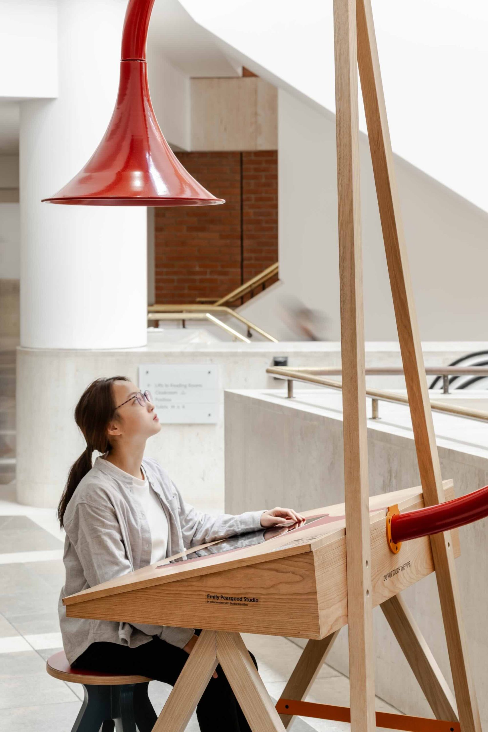 Listening Desk by Emily Peasgood is a mid-century-inspired desk with a very large red metallic horn joined to it. It emerges out the back and round all the way over the desk, finishing above the listener's head. From the side, it looks like a huge base clef or ear. It looks like something from Dr Seuss. Sound can only be heard from under the horn. There is bespoke software on the screen that allows members of the public to listen to archival sound recordings and remix them. This is a close-up of a woman sitting at the desk. She is looking up at the horn. She wears a white shirt, glasses and has brown hair in a ponytail.