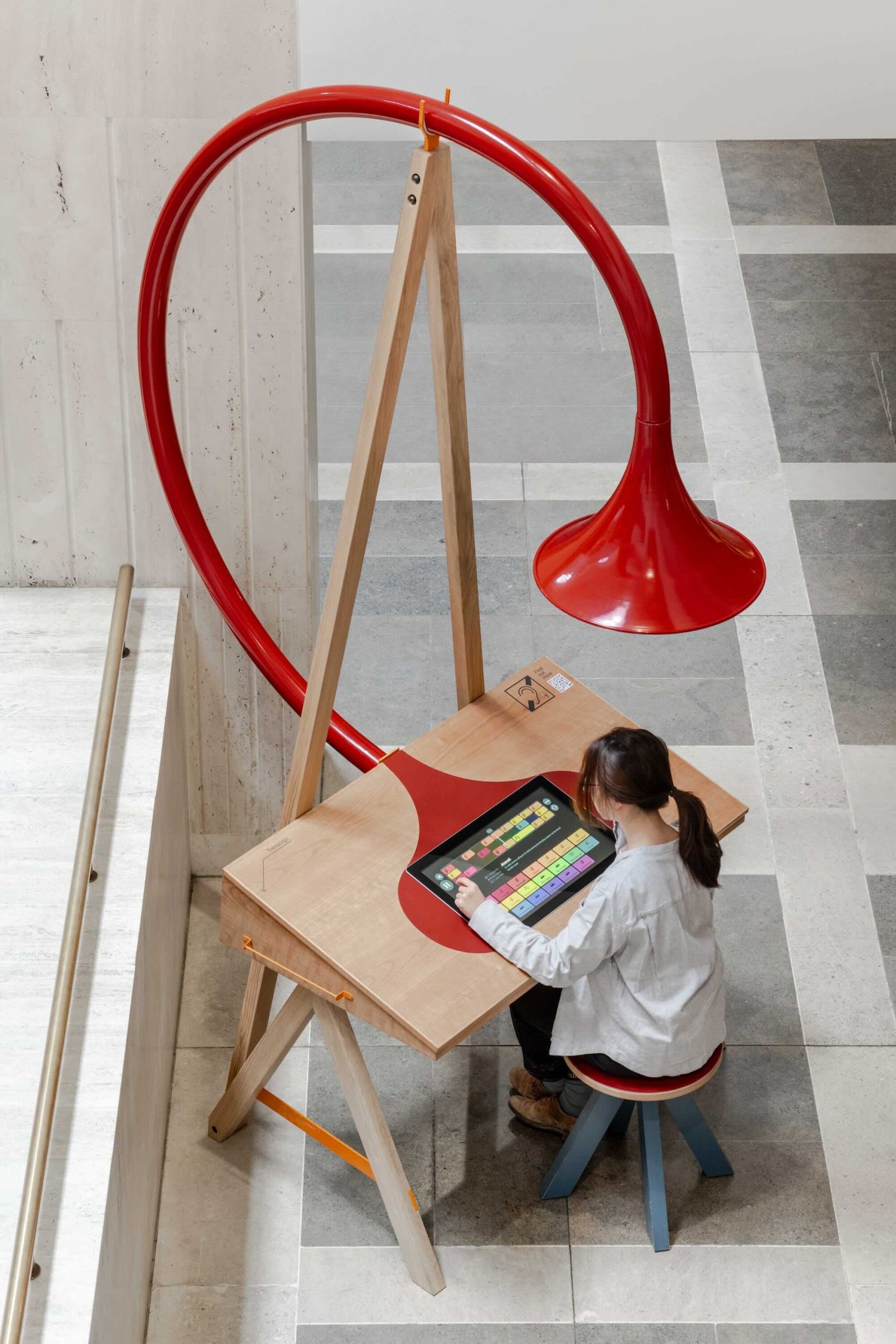 Listening Desk by Emily Peasgood is a mid-century-inspired desk with a very large red metallic horn joined to it. It emerges out the back and round all the way over the desk, finishing above the listener's head. From the side, it looks like a huge base clef or ear. It looks like something from Dr Seuss. Sound can only be heard from under the horn. There is bespoke software on the screen that allows members of the public to listen to archival sound recordings and remix them. This is a bird's-eye view of The Listening Desk from a higher floor at the British Library. A woman is sitting on the stool playing with the software on the touchscreen. She wears a white shirt and has a brown ponytail.
