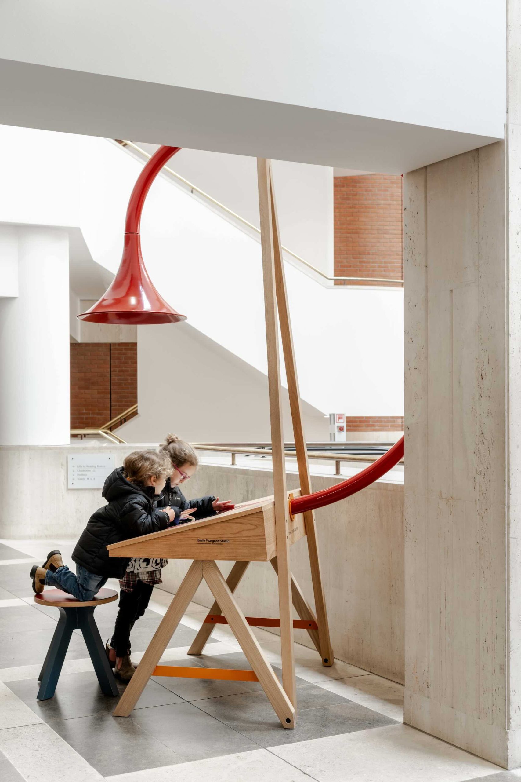 Listening Desk by Emily Peasgood is a mid-century-inspired desk with a very large red metallic horn joined to it. It emerges out the back and round all the way over the desk, finishing above the listener's head. From the side, it looks like a huge base clef or ear. It looks like something from Dr Seuss. Sound can only be heard from under the horn. There is bespoke software on the screen that allows members of the public to listen to archival sound recordings and remix them. This image shows two young children playing with the desk. One is kneeling on the stool while the other is standing next to them. Both are wearing blue jeans and have light brown hair.