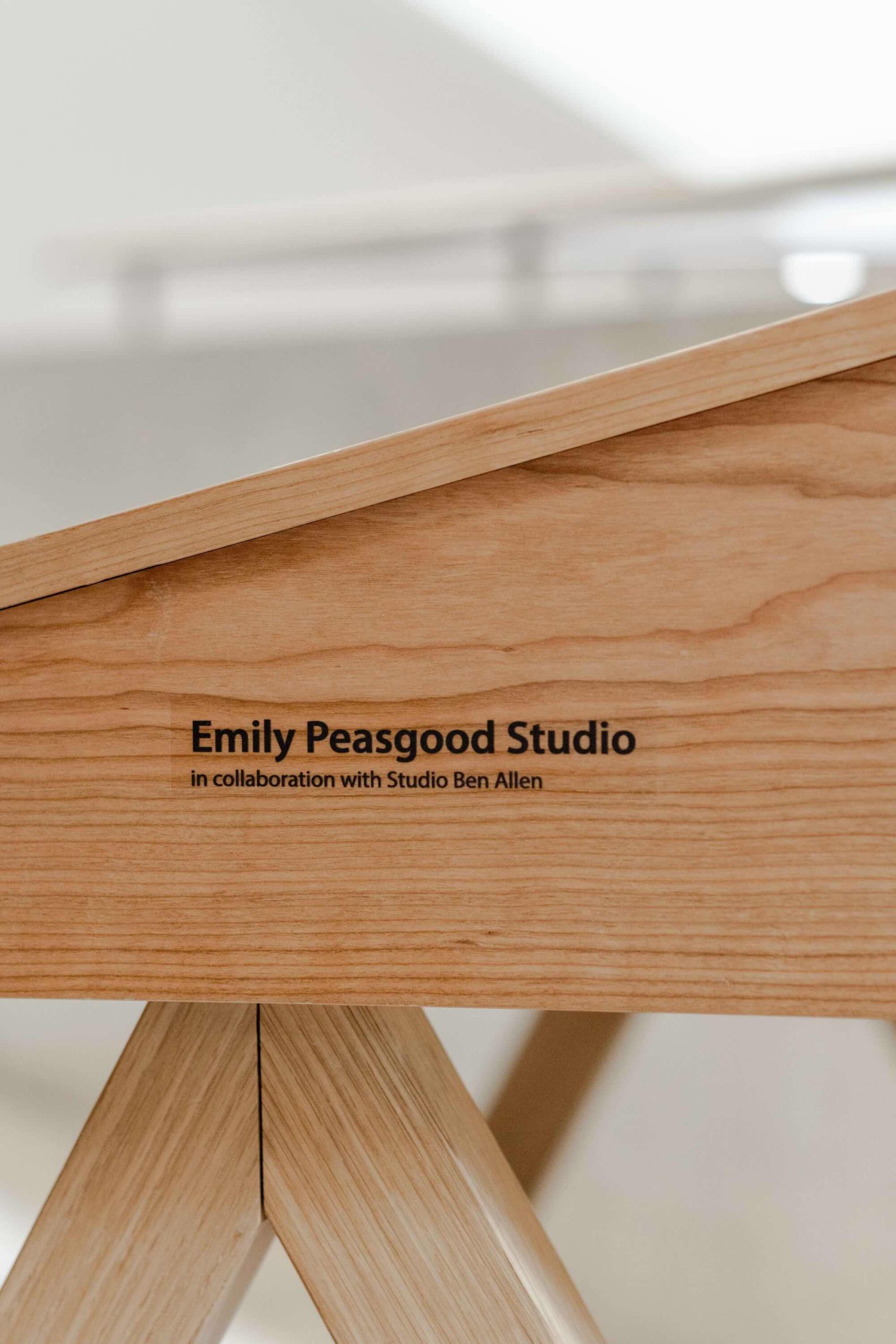 Listening Desk by Emily Peasgood is a mid-century-inspired desk with a very large red metallic horn joined to it. It emerges out the back and round all the way over the desk, finishing above the listener's head. From the side, it looks like a huge base clef or ear. It looks like something from Dr Seuss. Sound can only be heard from under the horn. There is bespoke software on the screen that allows members of the public to listen to archival sound recordings and remix them. This image shows the side profile of the desk top, with a view of the credit which states: 