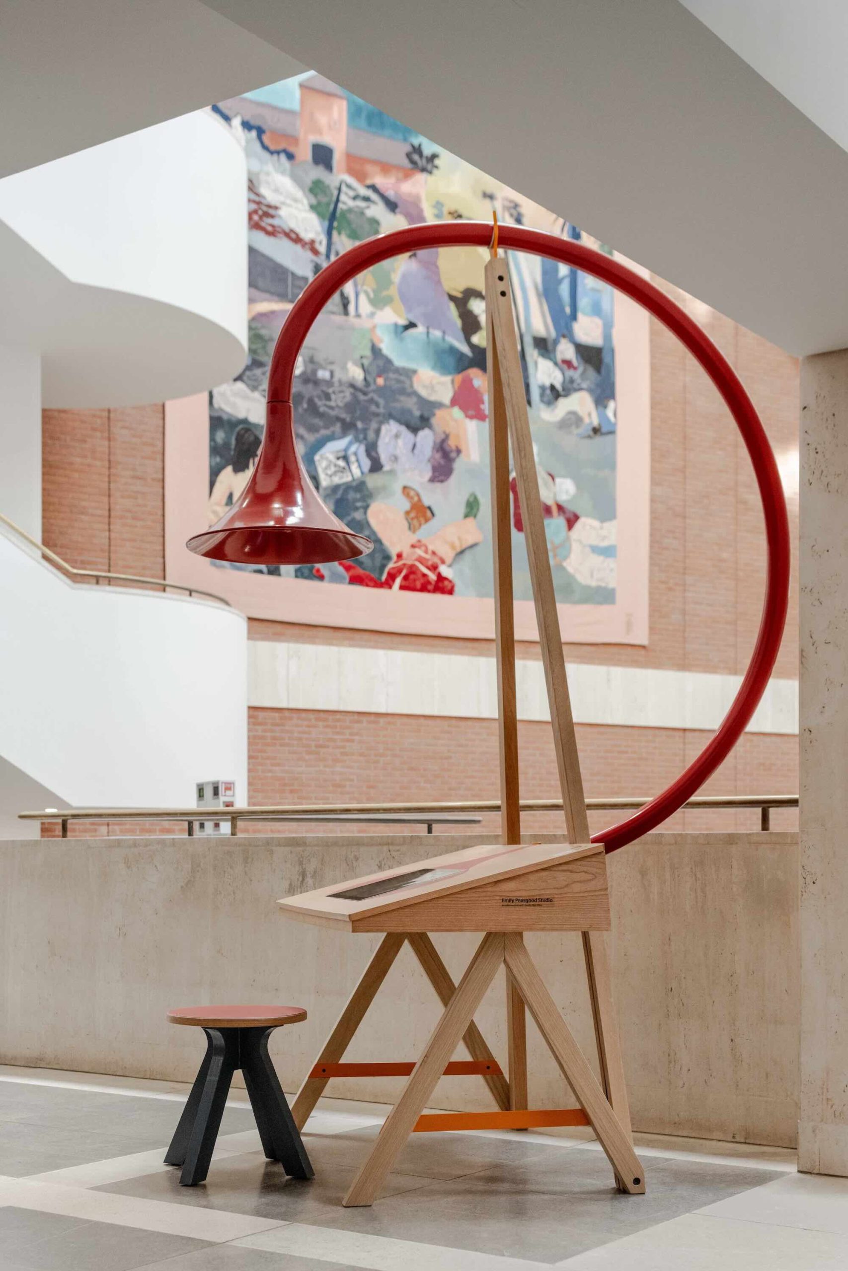 Listening Desk by Emily Peasgood is a mid-century-inspired desk with a very large red metallic horn joined to it. It emerges out the back and round all the way over the desk, finishing above the listener's head. From the side, it looks like a huge base clef or ear. It looks like something from Dr Seuss. Sound can only be heard from under the horn. There is bespoke software on the screen that allows members of the public to listen to archival sound recordings and remix them. This is a side profile of the desk with a small red-topped stool placed underneath the horn.