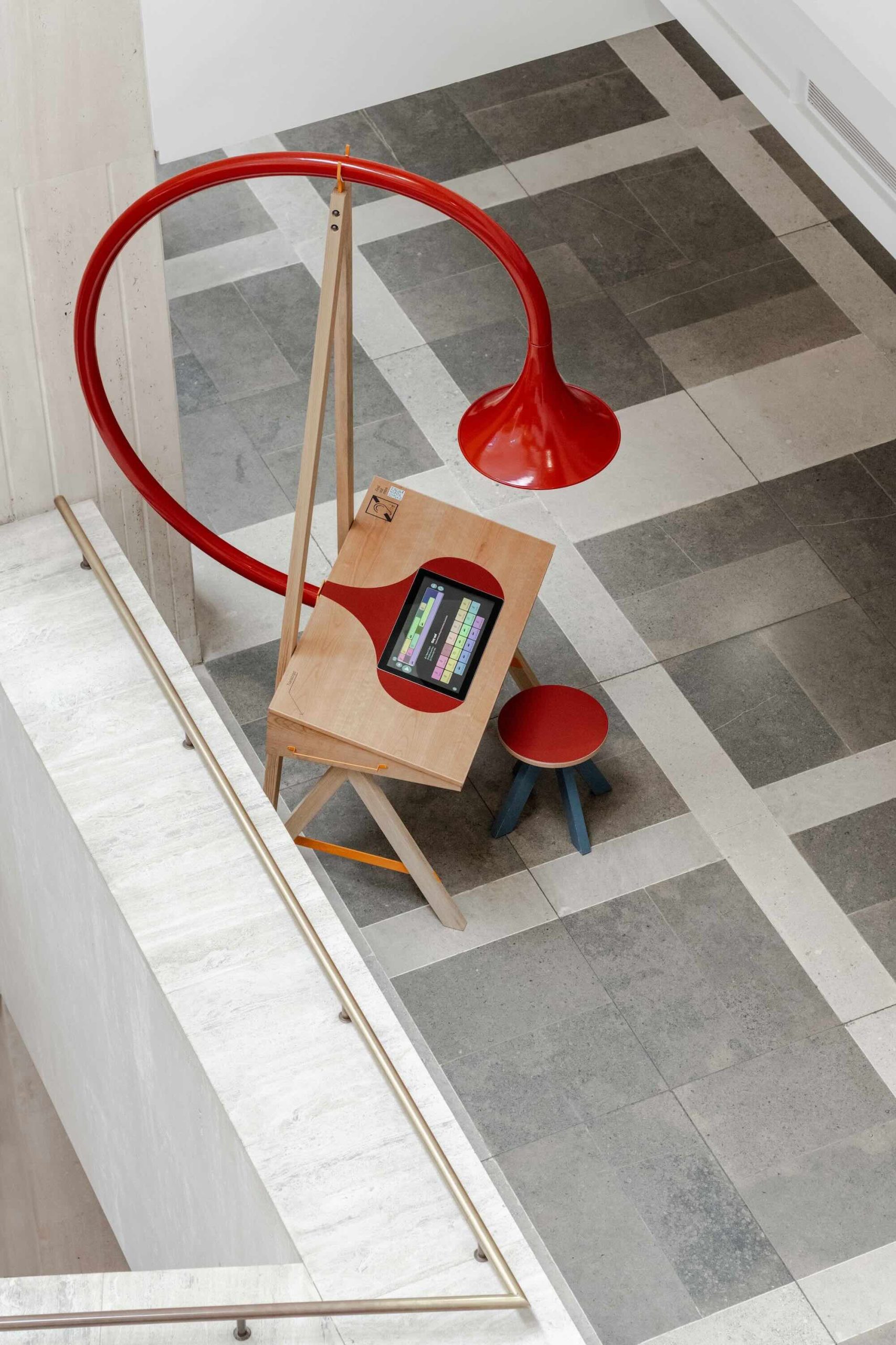 Listening Desk by Emily Peasgood is a mid-century-inspired desk with a very large red metallic horn joined to it. It emerges out the back and round all the way over the desk, finishing above the listener's head. From the side, it looks like a huge base clef or ear. It looks like something from Dr Seuss. Sound can only be heard from under the horn. There is bespoke software on the screen that allows members of the public to listen to archival sound recordings and remix them. This is a bird's-eye photograph of the desk taken from the floor above in the British Library.