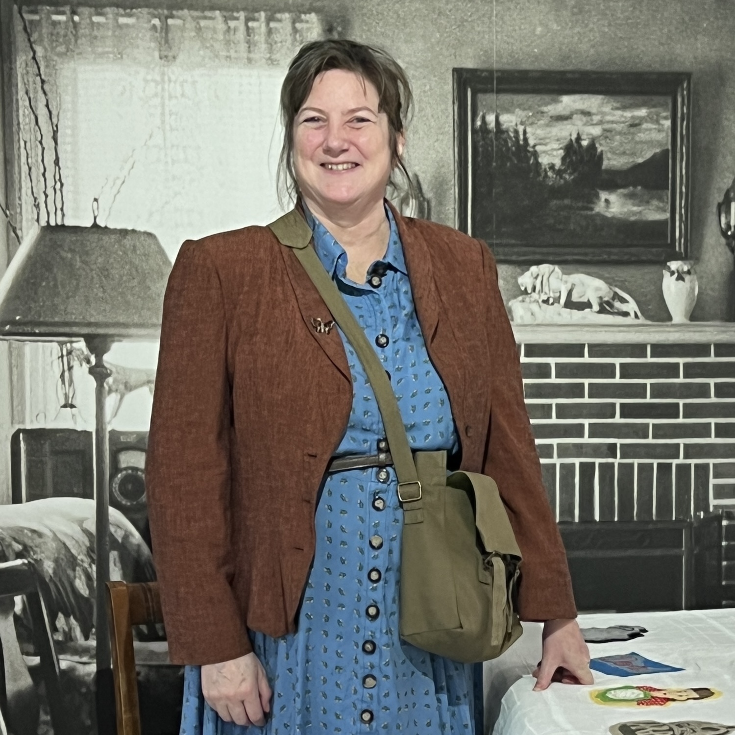 Jacquie_Ansell_Elmbridge This is a picture of Jacqui Ansell who sewed the patches onto the tablecloth and led the community sewing workshops. She is standing next to Threaded Shelter by Emily Peasgood and smiling at the camera. She has brown hair, a dark brown jacket, a blue dress and a green shoulder bag. She looks like a WWII land girl.