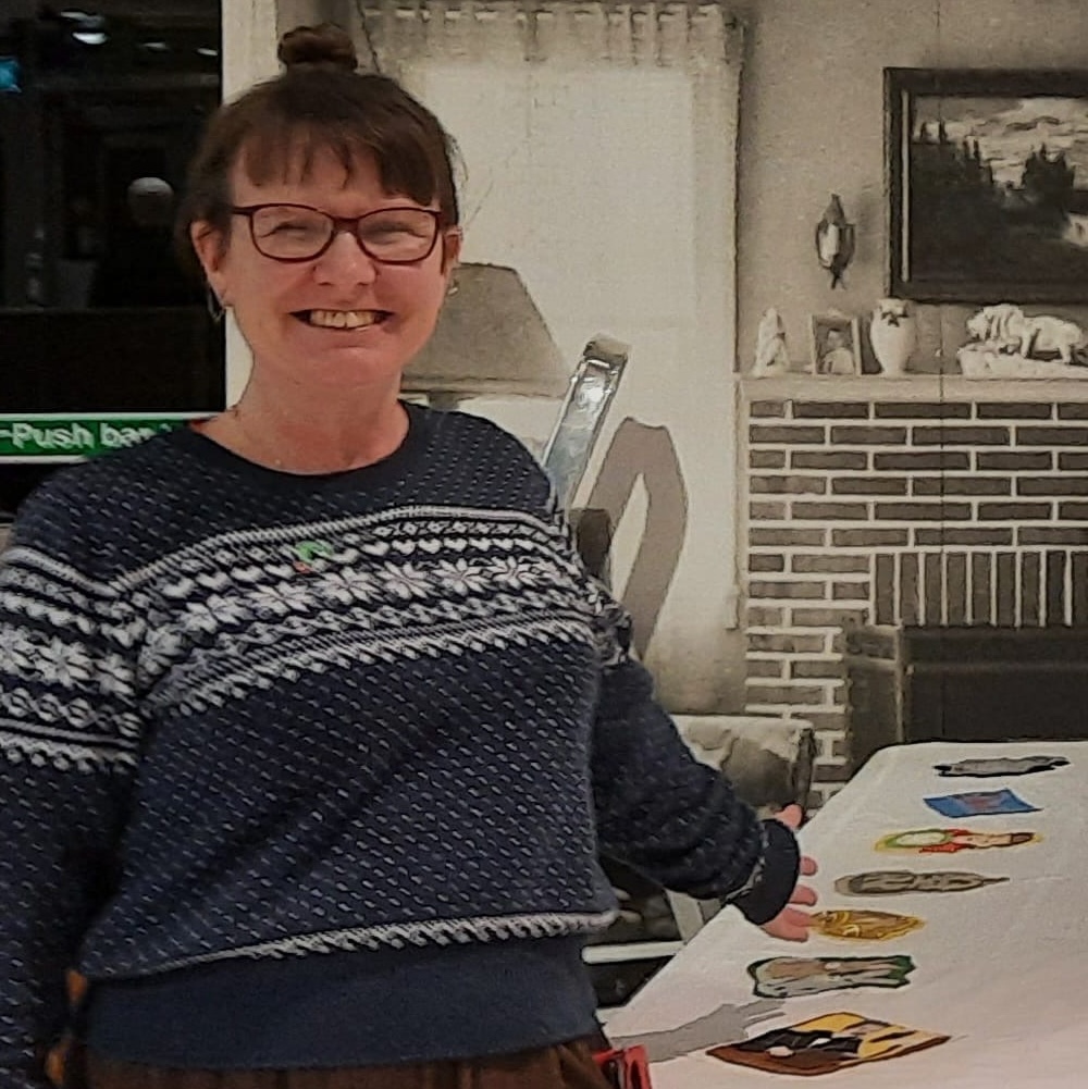 Kate_Lynn_Devere_Elmbridge This is a picture of Kate Lynn-Devere who was a workshop facilitator for the community sewing workshops. She has brown hair with a fringe, glasses and a navy blue jumper. She is standing in front of the tablecloth and pointing at it while smiling to the camera.