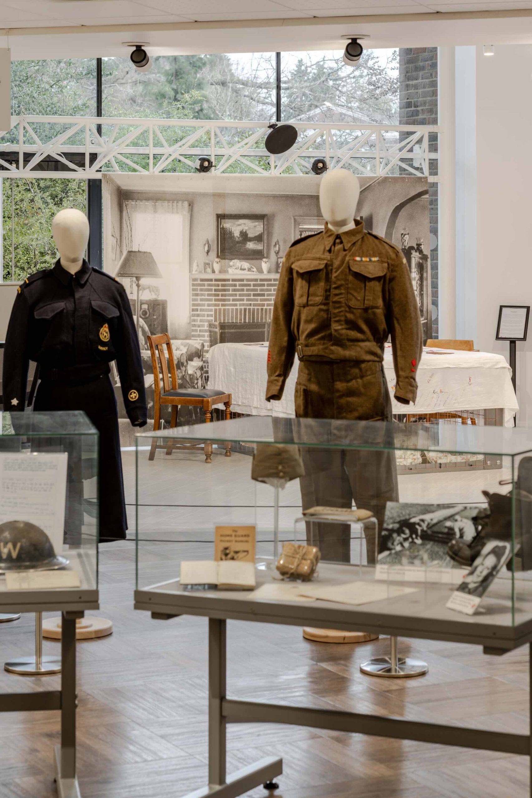 This is a photo of Threaded Shelter by Emily Peasgood. It is a recreation of a Morrison Shelter from WW3 with a bespoke tablecloth containing clothing repair patches. When you touch each patch, a voice tells a story. This photo shows the wider exhibition it was a part of: Echoes of War. In the forefront, there are two mannequins dressed in WWII uniforms, and two glass display cases showing a range of WWII ephemera, including an ARP helmet and a photograph in a frame. Threaded Shelter is further back, at the read of the space.