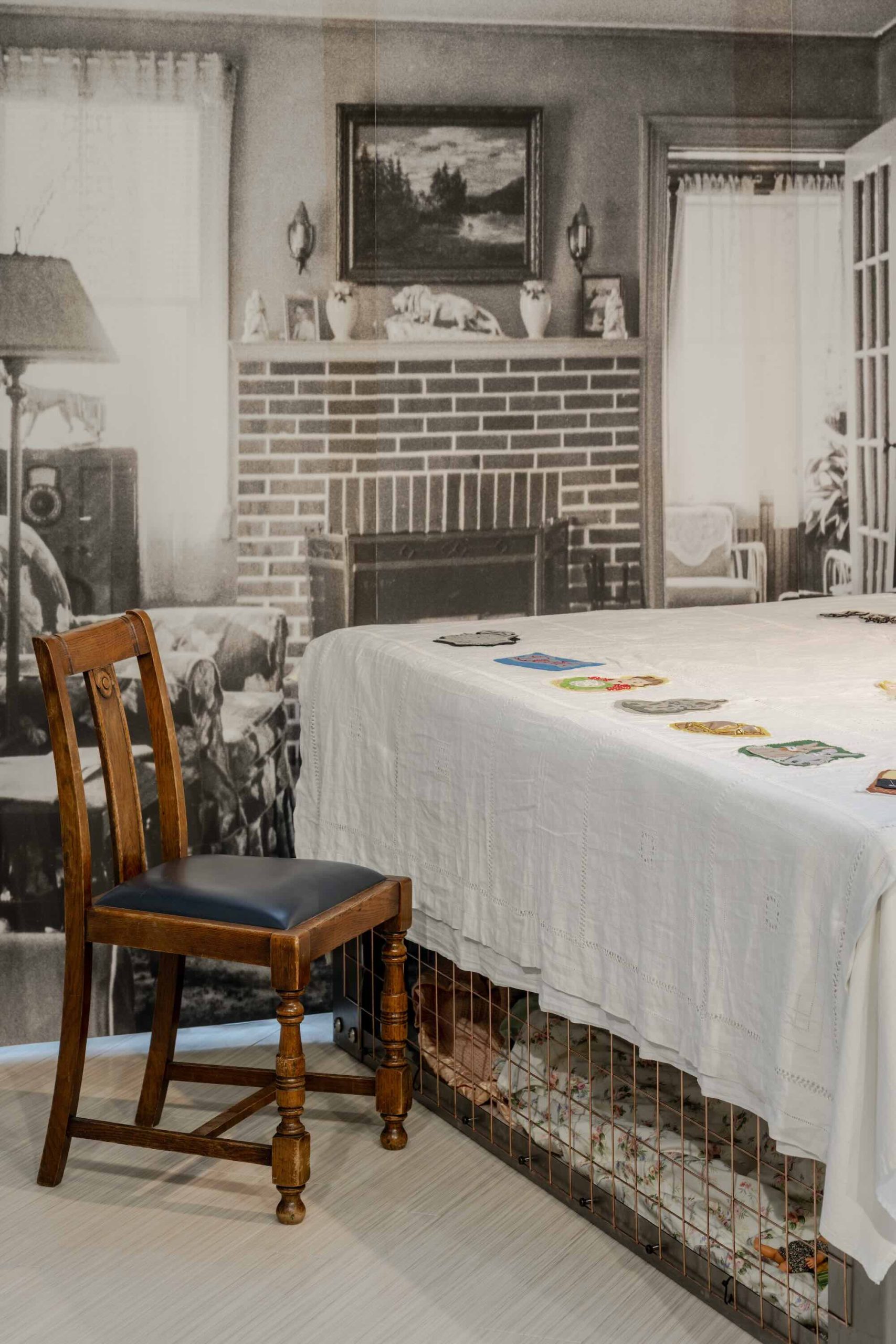 This is a photo of Threaded Shelter by Emily Peasgood. It is a recreation of a Morrison Shelter from WW3 with a bespoke tablecloth containing clothing repair patches. When you touch each patch, a voice tells a story. This is a side view of the shelter with a good view of the WWII living room backdrop and a dining chair with a blue leather seat.