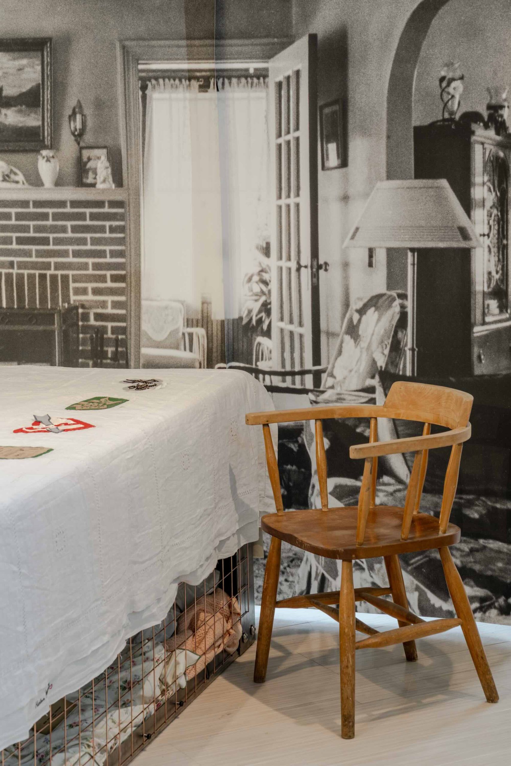 This is a photo of Threaded Shelter by Emily Peasgood. It is a recreation of a Morrison Shelter from WW3 with a bespoke tablecloth containing clothing repair patches. When you touch each patch, a voice tells a story. This photo shows a side view of the table with a good image of the backdrop showing a typical WWII living room in black and white and an antique wooden captain's chair.