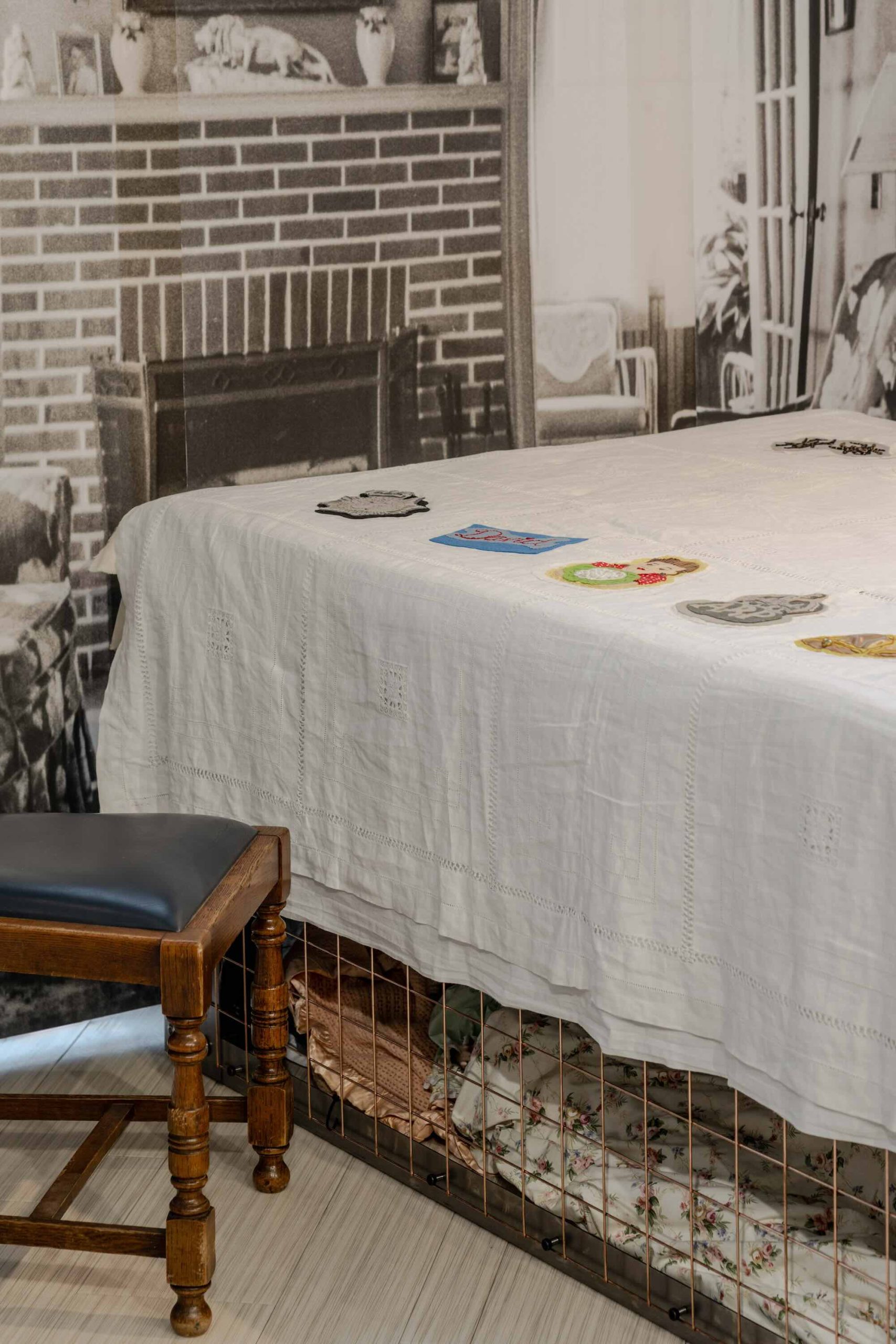 This is a photo of Threaded Shelter by Emily Peasgood. It is a recreation of a Morrison Shelter from WW3 with a bespoke tablecloth containing clothing repair patches. When you touch each patch, a voice tells a story. This photo is a side view showing some of the background, primarily a WWII fireplace, and a dining chair with a leather blue seat cover.