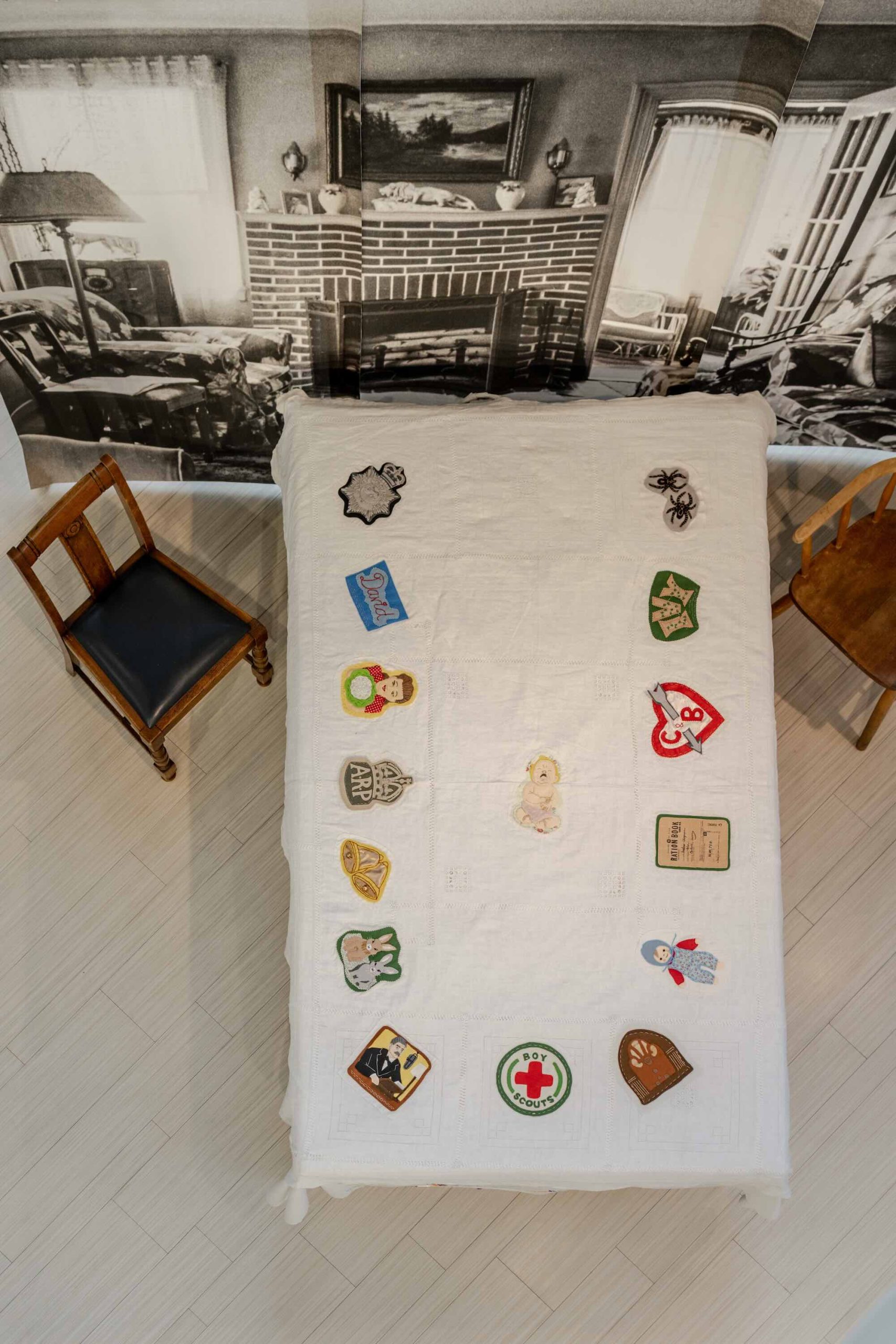 This is a photo of Threaded Shelter by Emily Peasgood. It is a recreation of a Morrison Shelter from WW3 with a bespoke tablecloth containing clothing repair patches. When you touch each patch, a voice tells a story. This photo is a bird's-eye view of the table. Looking down from above, there are 16 patches.