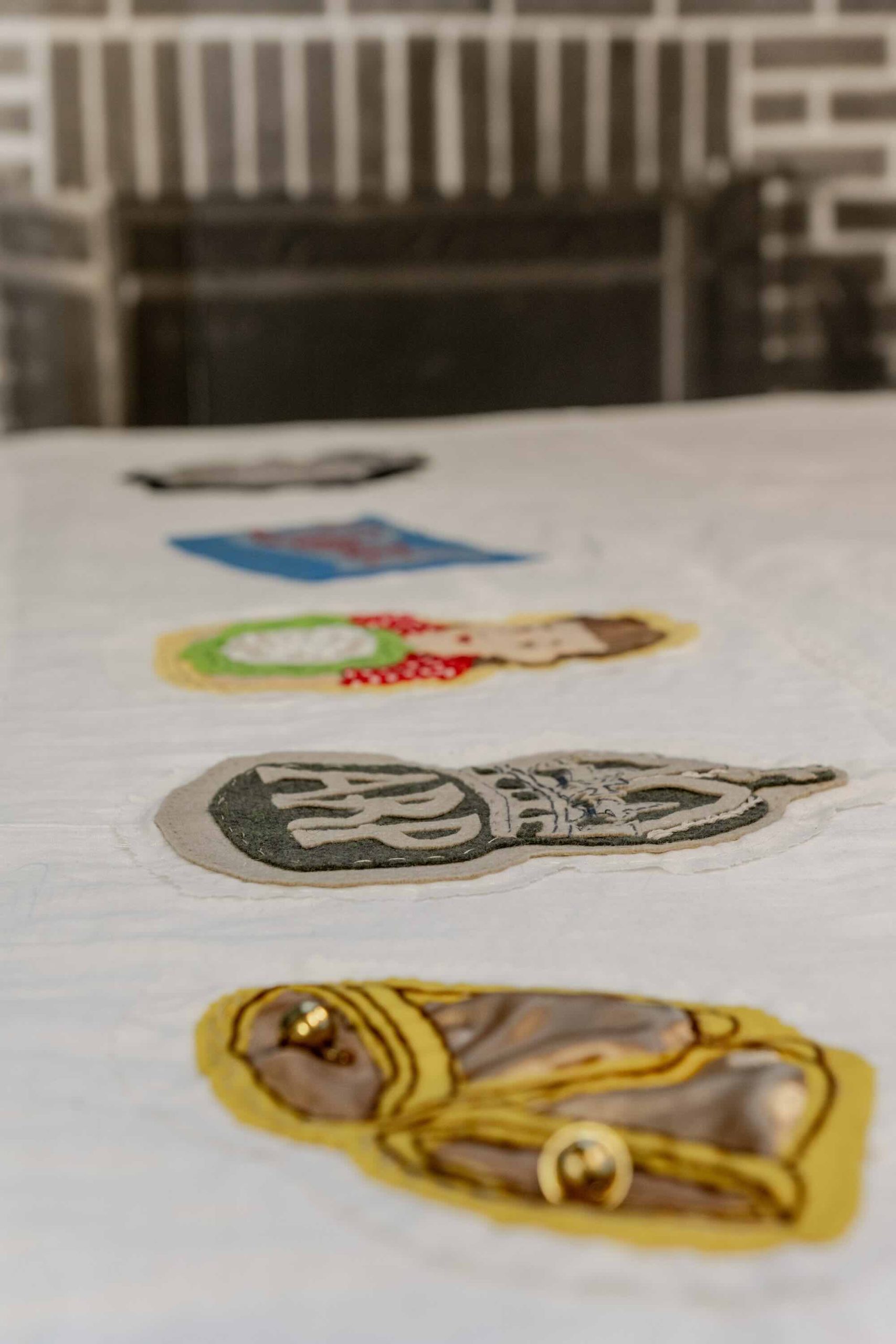 This is a photo of Threaded Shelter by Emily Peasgood. It is a recreation of a Morrison Shelter from WW3 with a bespoke tablecloth containing clothing repair patches. When you touch each patch, a voice tells a story. This photo focuses on a patch of two bells, and an ARP patch.
