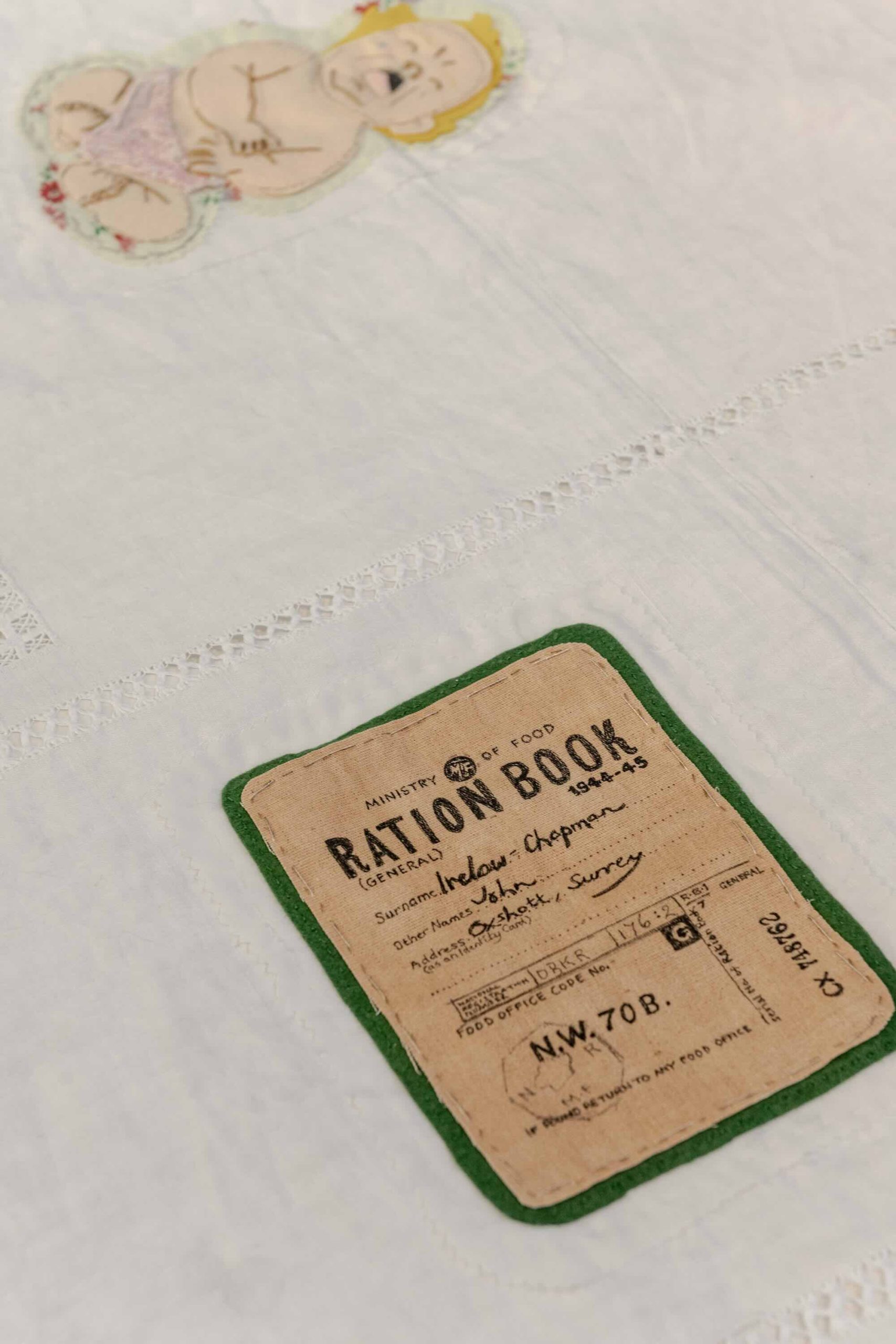 This is a photo of Threaded Shelter by Emily Peasgood. It is a recreation of a Morrison Shelter from WW3 with a bespoke tablecloth containing clothing repair patches. When you touch each patch, a voice tells a story. This photo focuses on the Ration Book patch, and a patch of a crying baby in a pink nappy.
