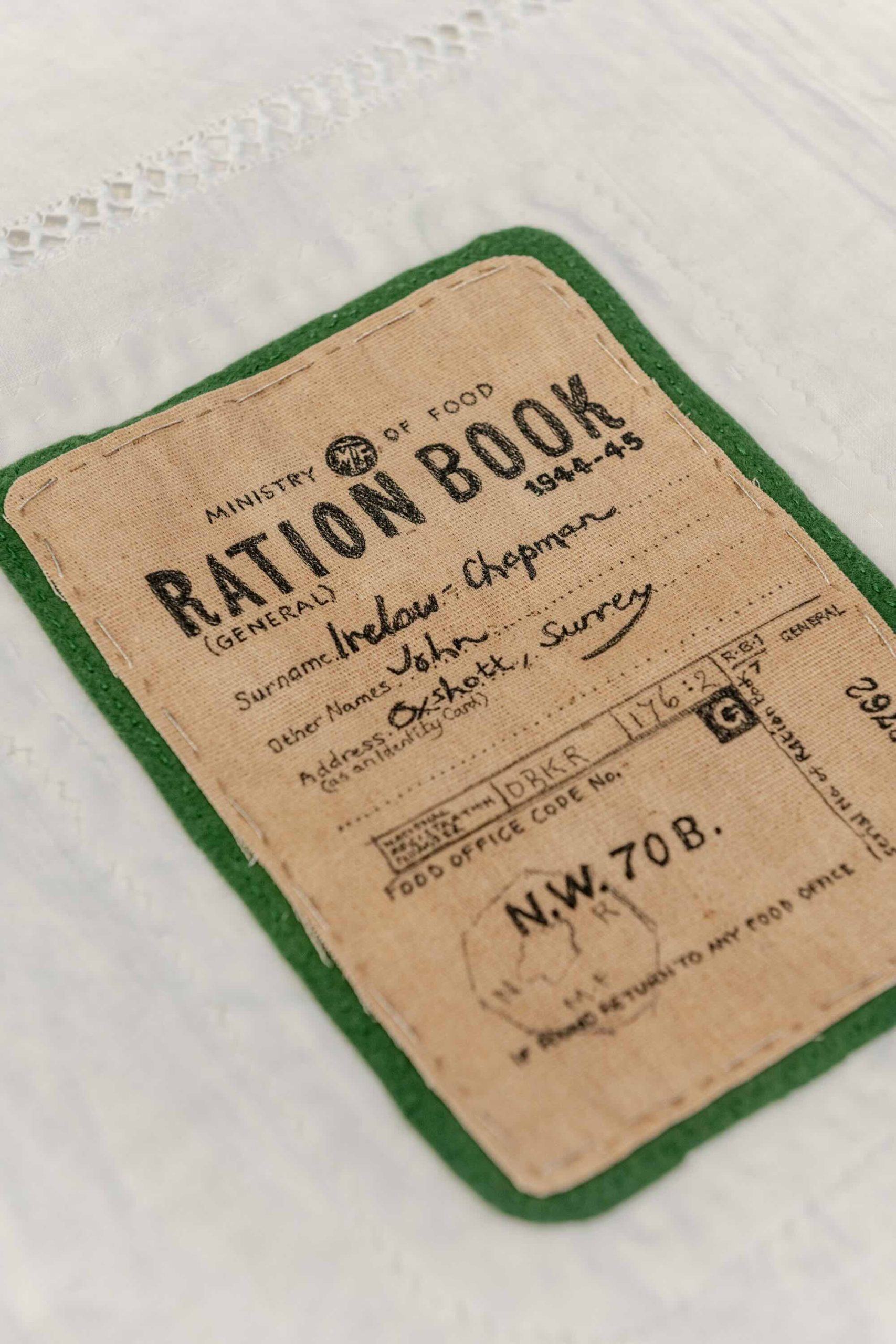 This is a photo of Threaded Shelter by Emily Peasgood. It is a recreation of a Morrison Shelter from WW3 with a bespoke tablecloth containing clothing repair patches. When you touch each patch, a voice tells a story. This photo is a close-up image of a Ration Book patch, which includes the name John Irelow-Chapman, Oxshott, Surrey. It is very realistic-looking. The book is sewn onto a piece of brown hessian fabric, and there is green backing fabric around it, creating a border.