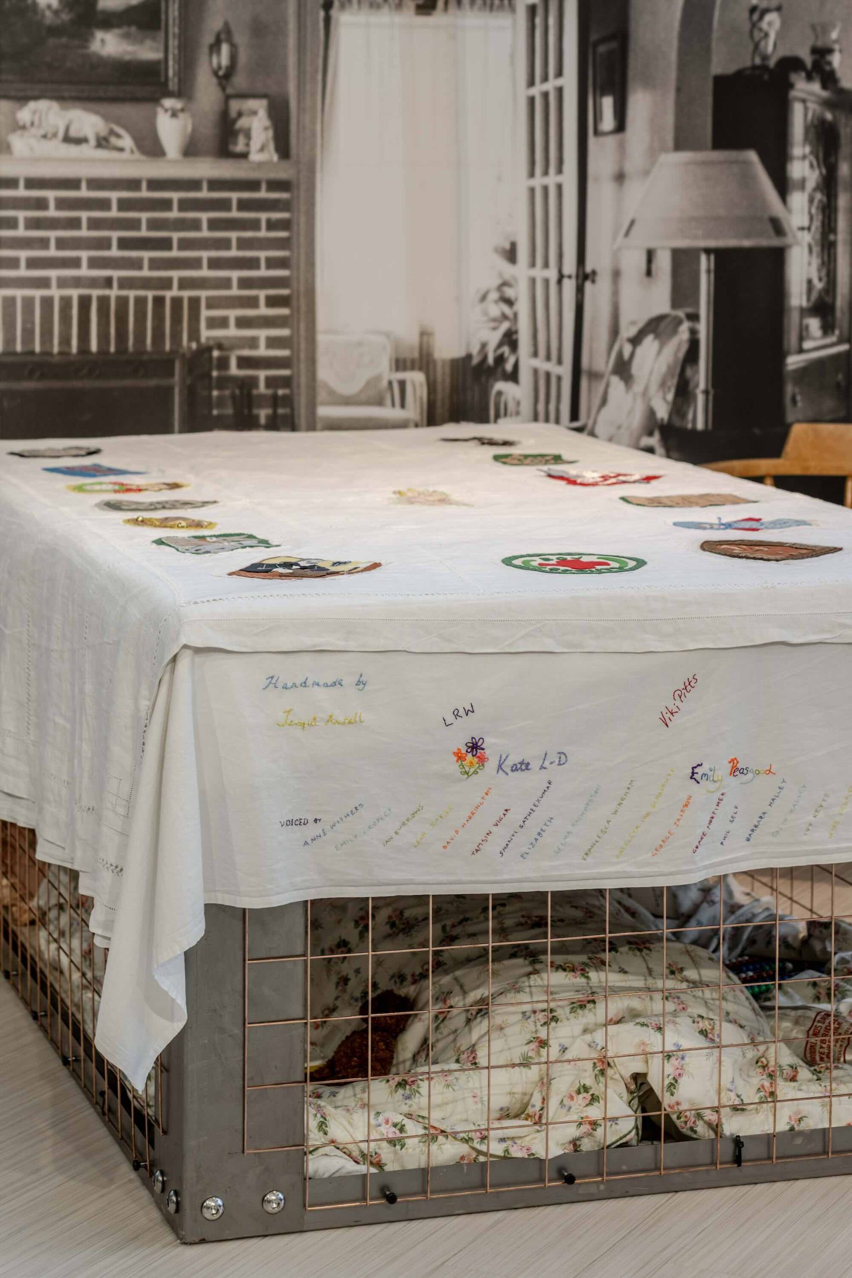 This is a photo of Threaded Shelter by Emily Peasgood. It is a recreation of a Morrison Shelter from WW3 with a bespoke tablecloth containing clothing repair patches. When you touch each patch, a voice tells a story.
