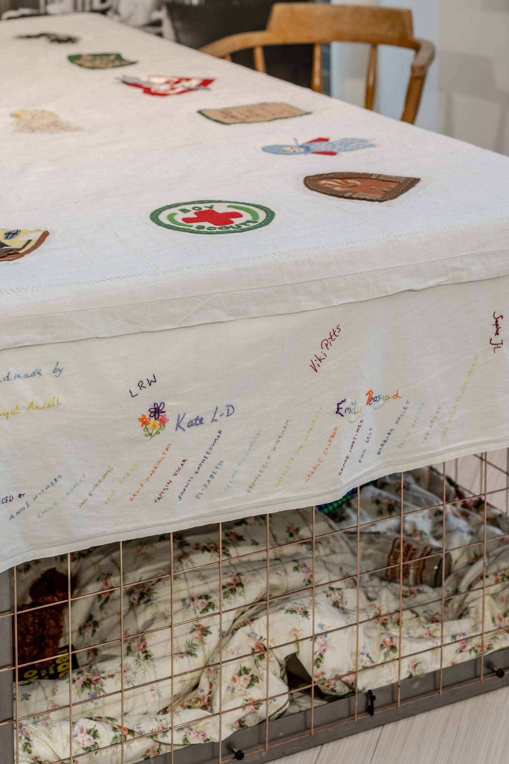 This is a photo of Threaded Shelter by Emily Peasgood. It is a recreation of a Morrison Shelter from WW3 with a bespoke tablecloth containing clothing repair patches. When you touch each patch, a voice tells a story. In this photo you can see the tablecloth, particularly the end panel which includes the stitched names of everyone who took part in the project. Lots of community members took part in recording stories and sewing the patches onto the tablecloth.