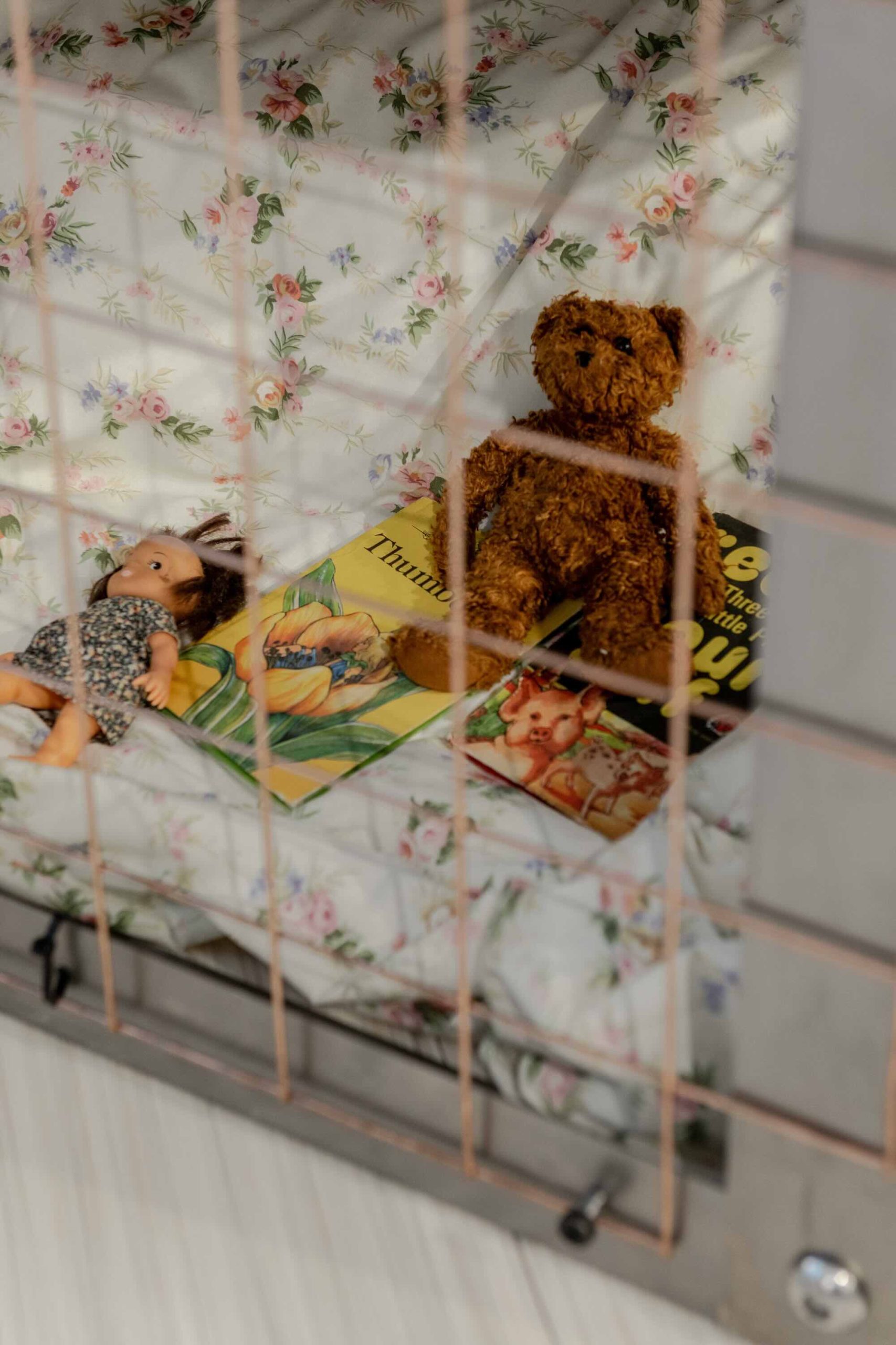 This is a photo of Threaded Shelter by Emily Peasgood. It is a recreation of a Morrison Shelter from WW3 with a bespoke tablecloth containing clothing repair patches. When you touch each patch, a voice tells a story. In this photo we see into the Morrison Shelter. There is some bedding and some children's toys: A small dolly with a flowery dress and brown hair, a small brown traditional teddy bear, and two vintage Ladybird books: Thumbelina and a reading book (title unclear).