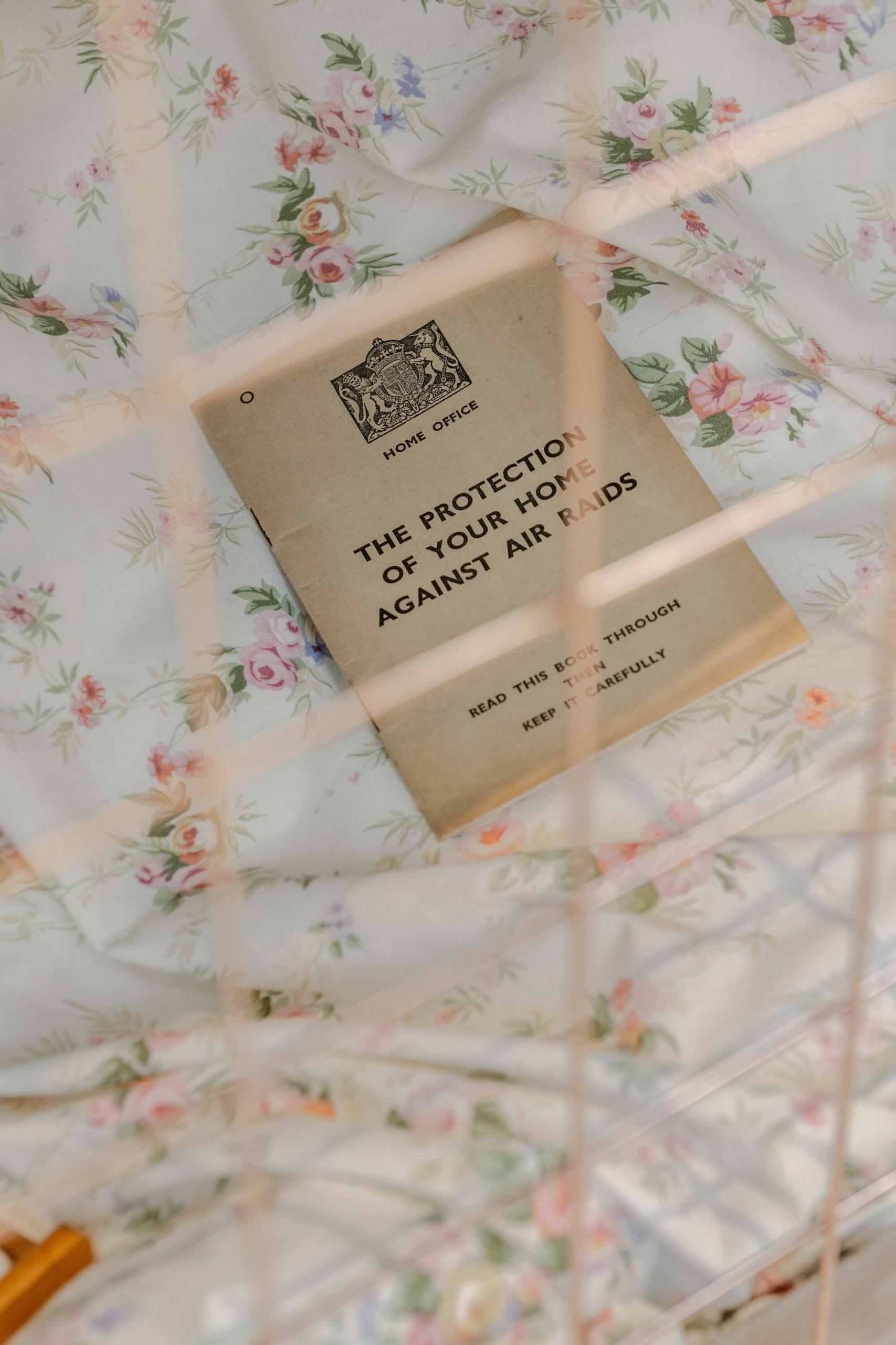 This is a photo of Threaded Shelter by Emily Peasgood. It is a recreation of a Morrison Shelter from WW3 with a bespoke tablecloth containing clothing repair patches. When you touch each patch, a voice tells a story. This photo shows inside the Morrison Shelter - you can see through the bars of the cage-like walls and onto some bedding where there is a booklet placed on top titled 'The Protection of Your Home Against Air Raids' - The Home Office.