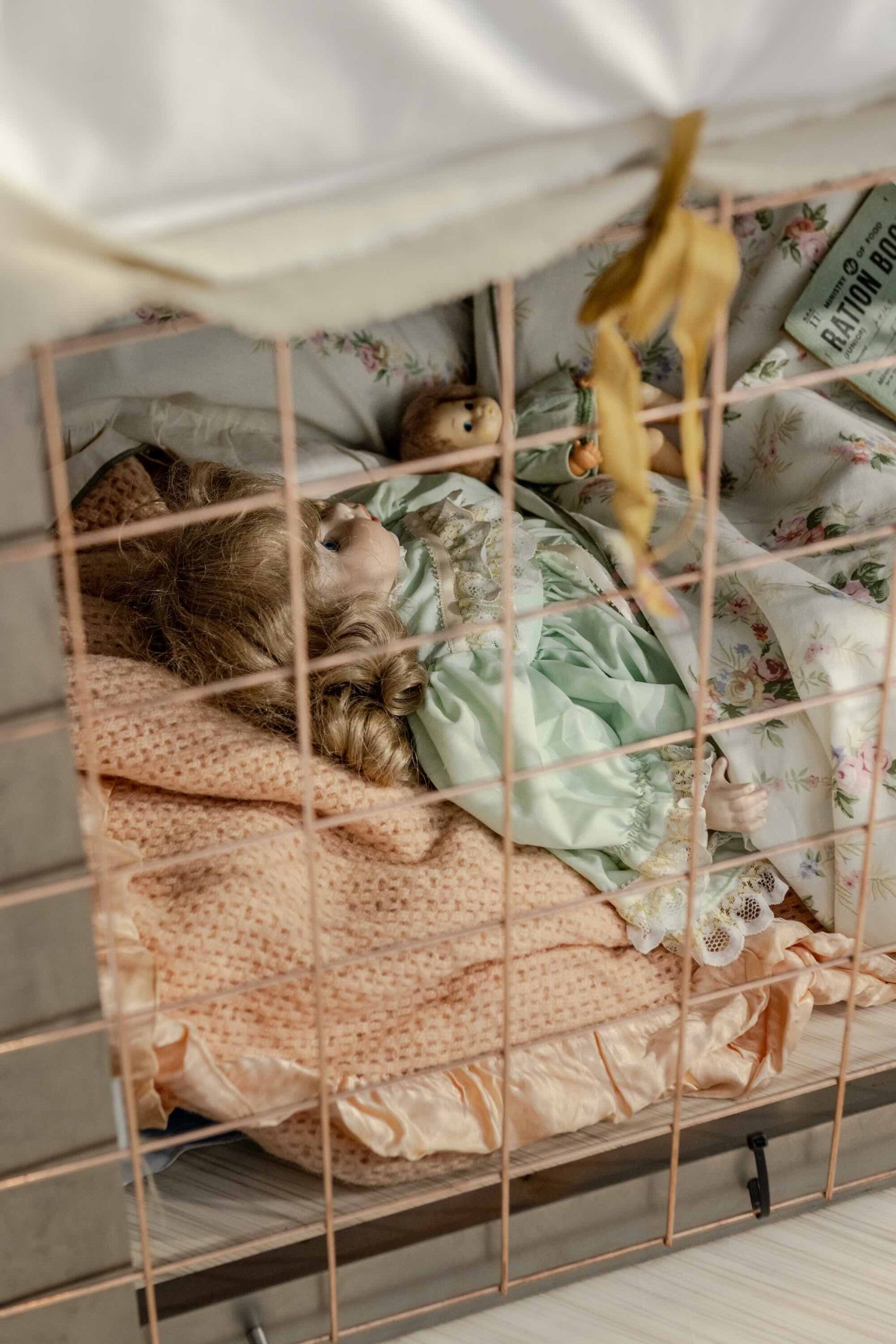 This is a photo of Threaded Shelter by Emily Peasgood. It is a recreation of a Morrison Shelter from WW3 with a bespoke tablecloth containing clothing repair patches. When you touch each patch, a voice tells a story. In this photo we see into the Morrison Shelter and there is some bedding and two children's dolly's placed inside. They are both wearing green dresses. One is a larger porcelin doll and the other is much smaller.