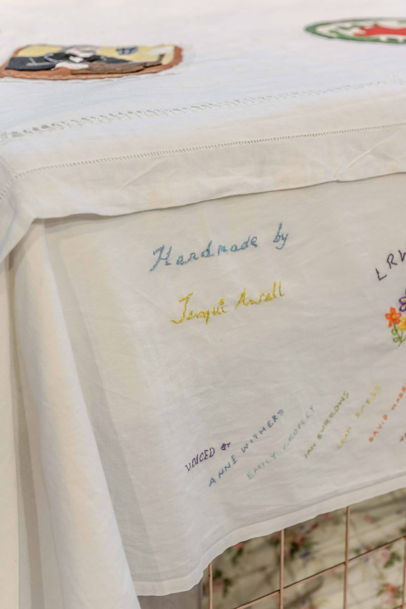 This is a photo of Threaded Shelter by Emily Peasgood. It is a recreation of a Morrison Shelter from WW3 with a bespoke tablecloth containing clothing repair patches. When you touch each patch, a voice tells a story. In this photo you can see the tablecloth, particularly the end panel which includes the stitched names of everyone who took part in the project. Lots of community members took part in recording stories and sewing the patches onto the tablecloth. The photo is quite zoomed in on 'Handmade by Jacqui Ansell' - Jacqui is a team member who sewed the patches and tablecloth and led the workshops with community members.